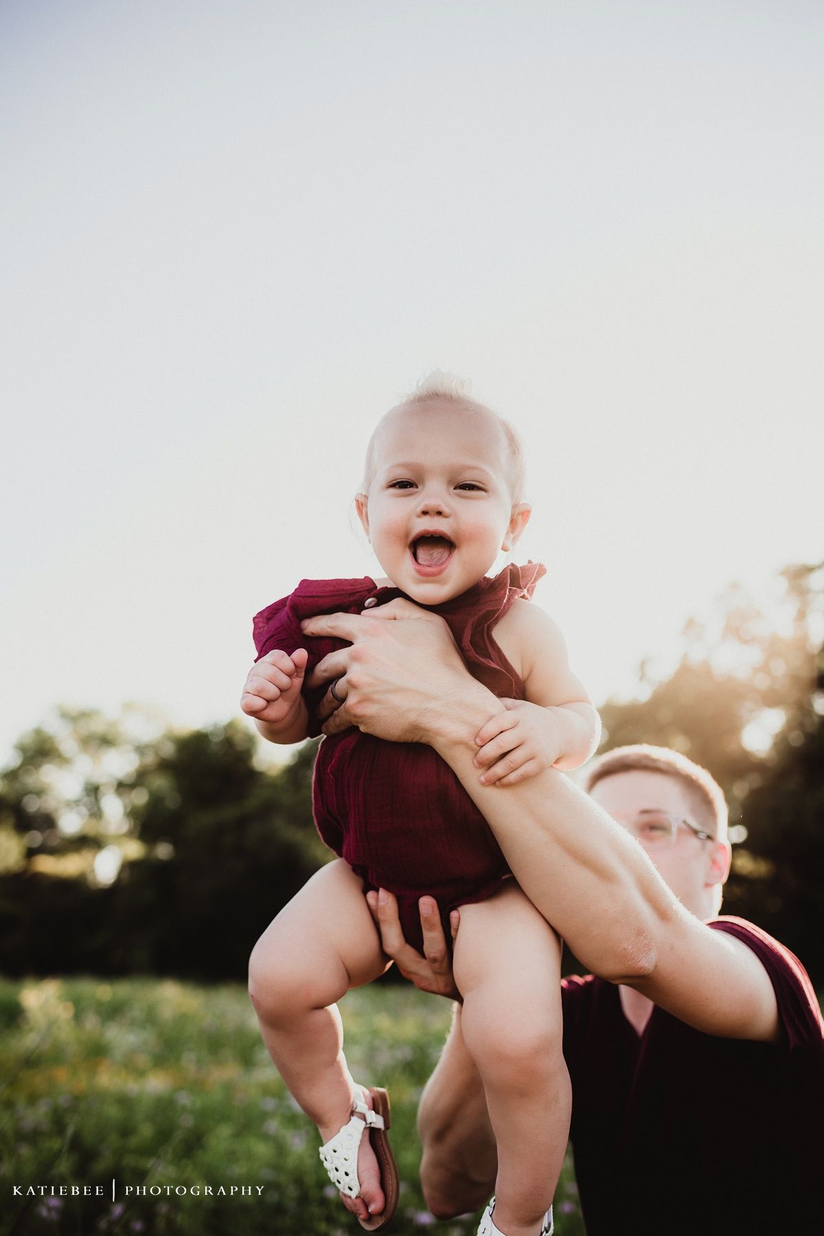 Candid moment of baby laughing while being lifted up against bright sky.