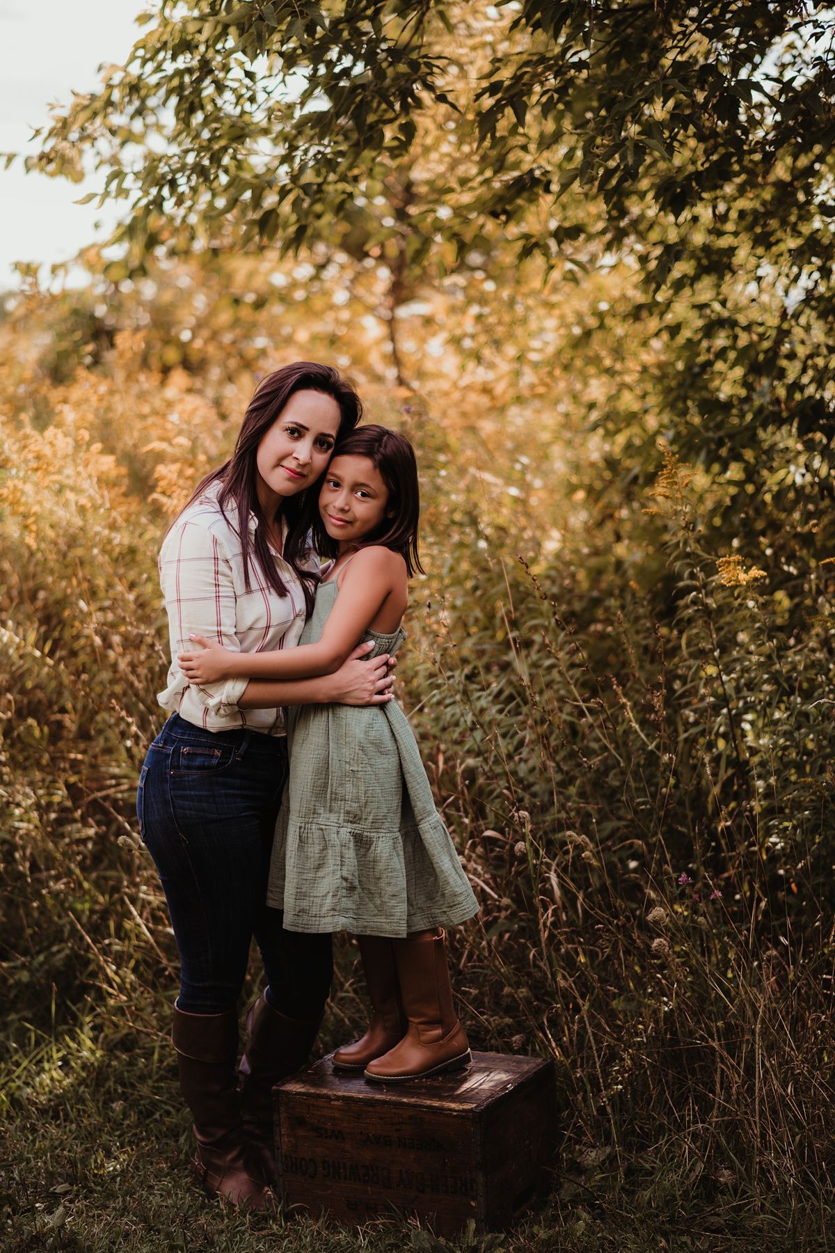 Two people embracing outdoors in golden sunlight while standing on a wooden box among autumn foliage.