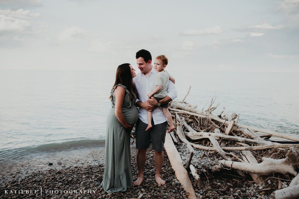 Family shares an intimate moment on a beach with driftwood and serene waters behind them.