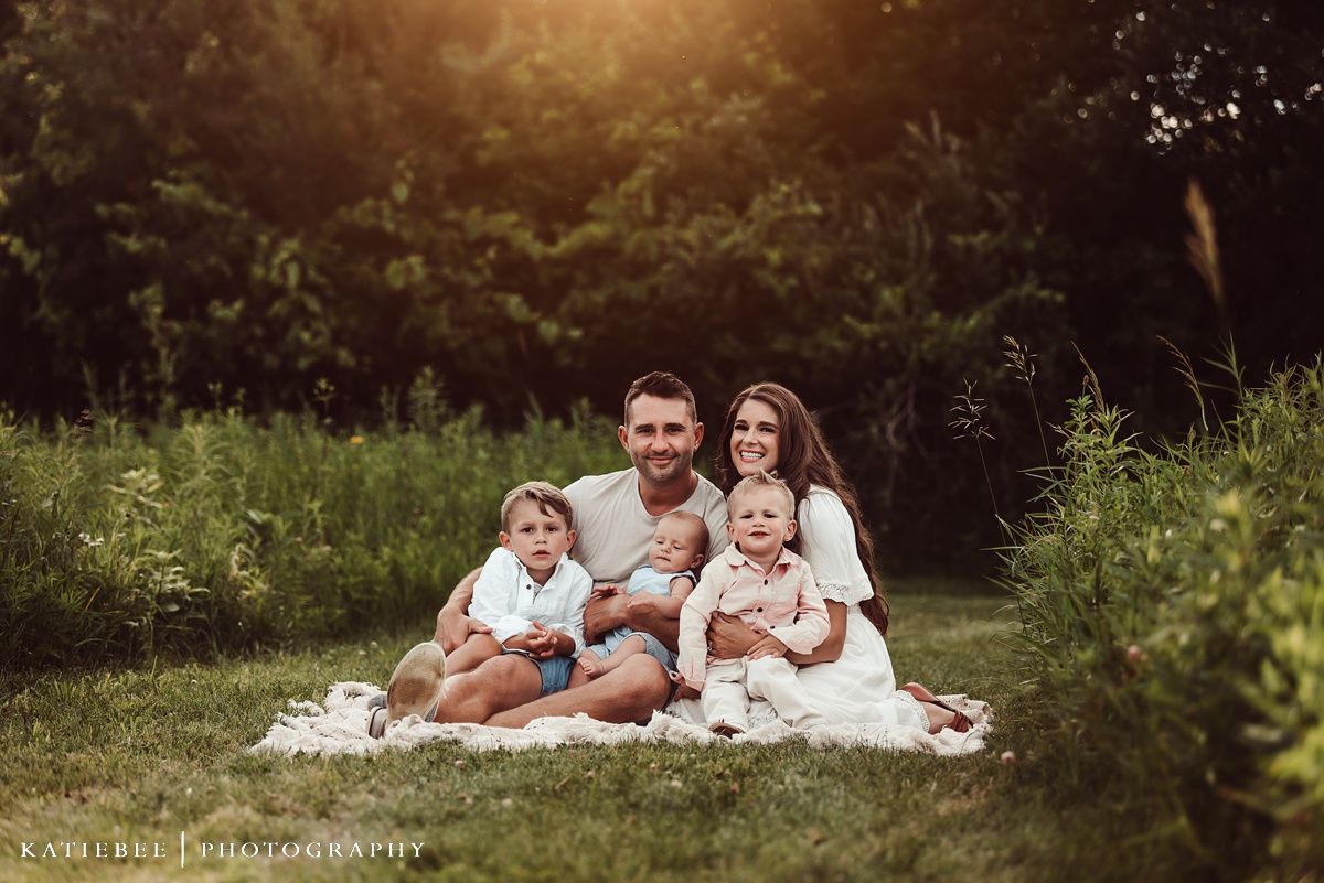 Family group portrait on white blanket in lush summer field during golden hour.