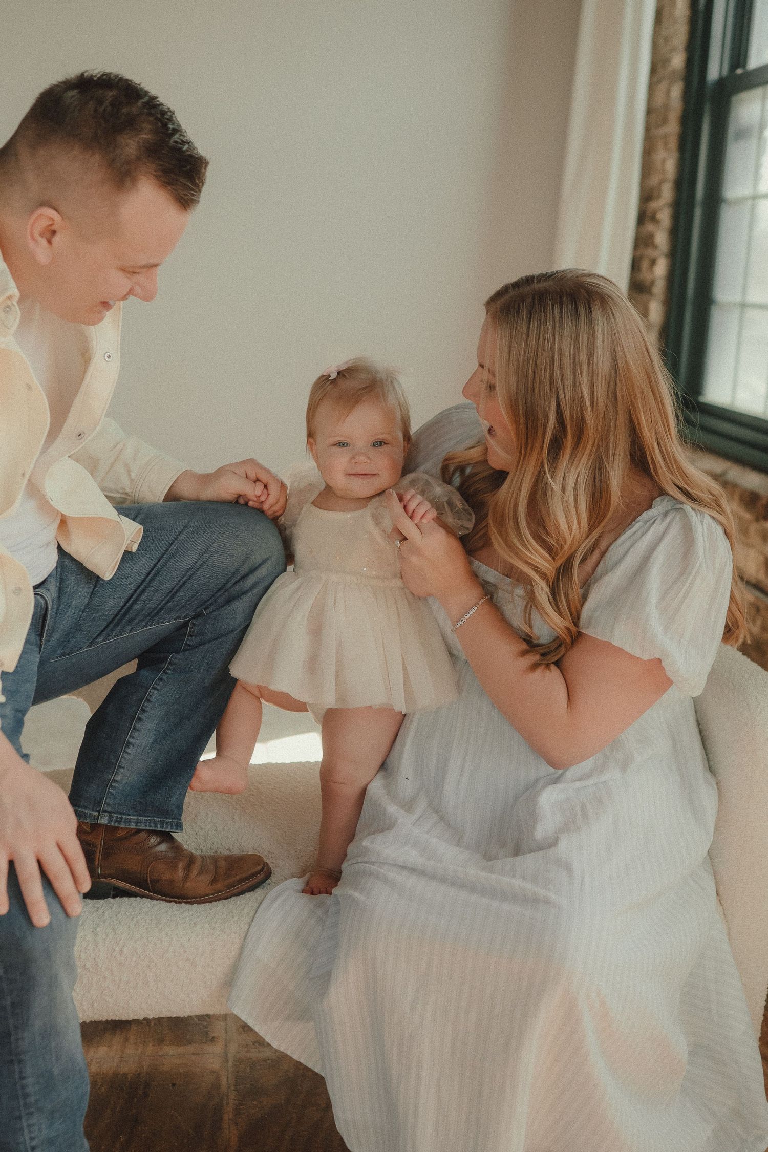 A young family with their baby girl in a cream dress share a tender moment together on a cozy white sofa.
