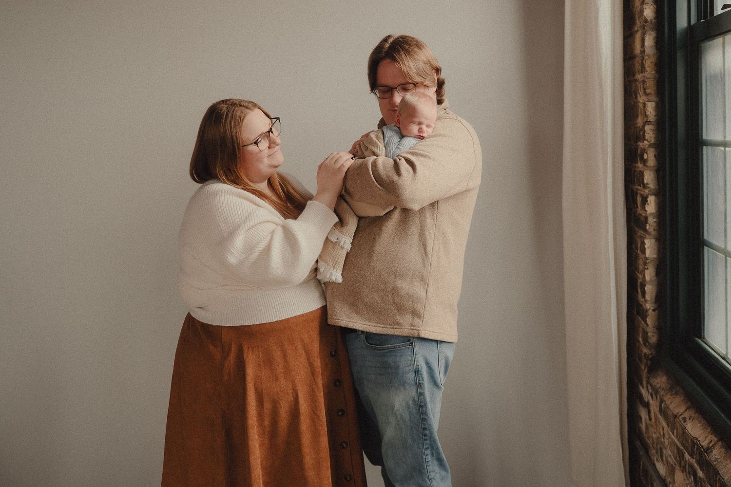A couple stands by a window holding their newborn baby, dressed in neutral tones, sharing a tender family moment.
