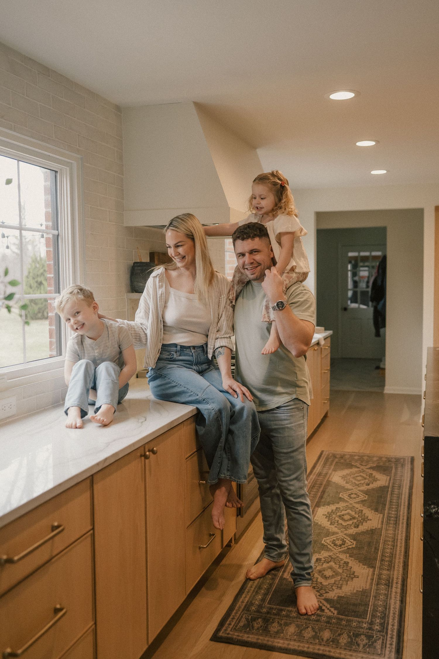 Happy family of four posing in a modern kitchen, parents sitting on counter with two young children, bright home interior.