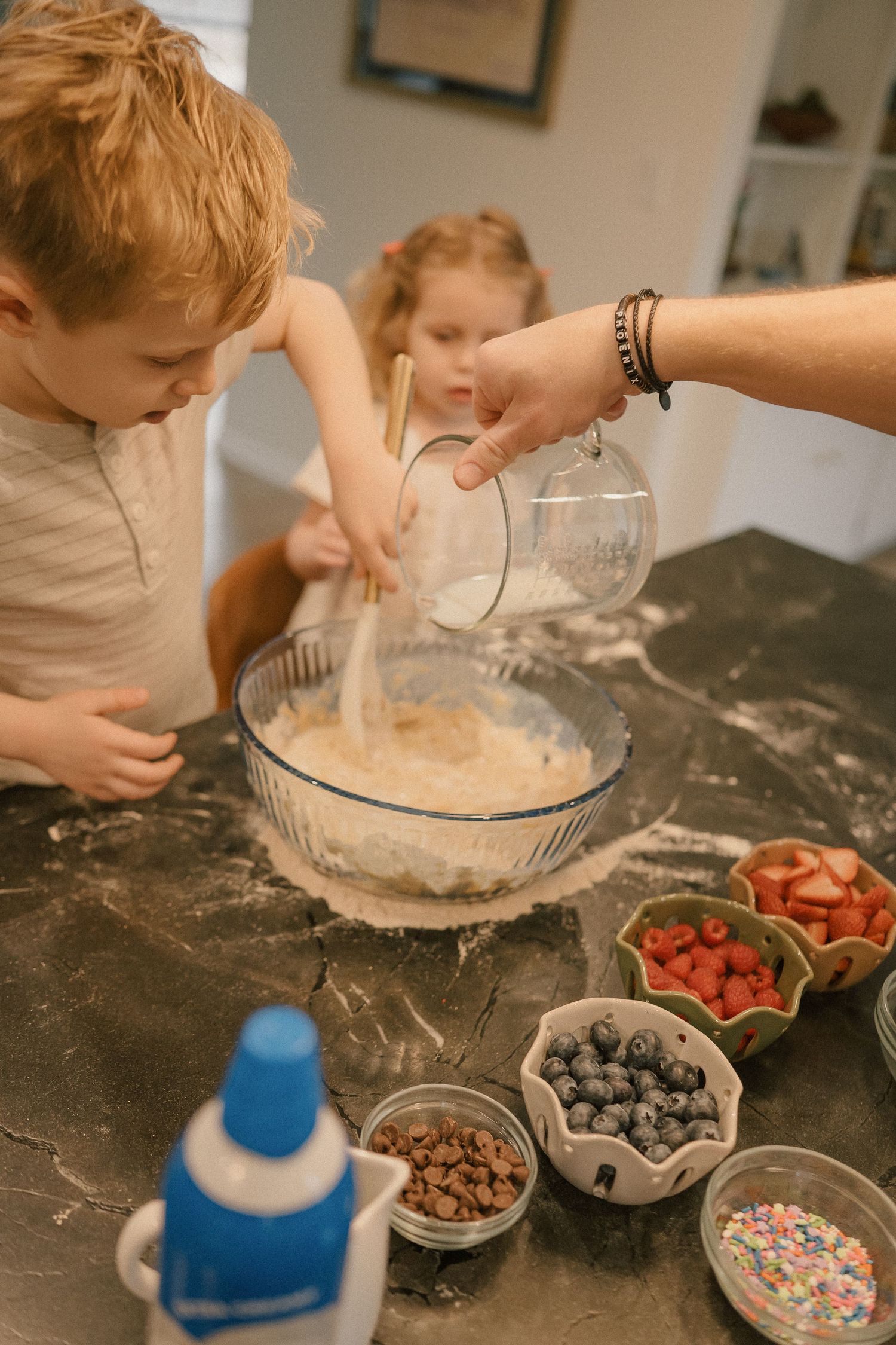 Children helping pour ingredients into mixing bowl with fresh berries on counter.
