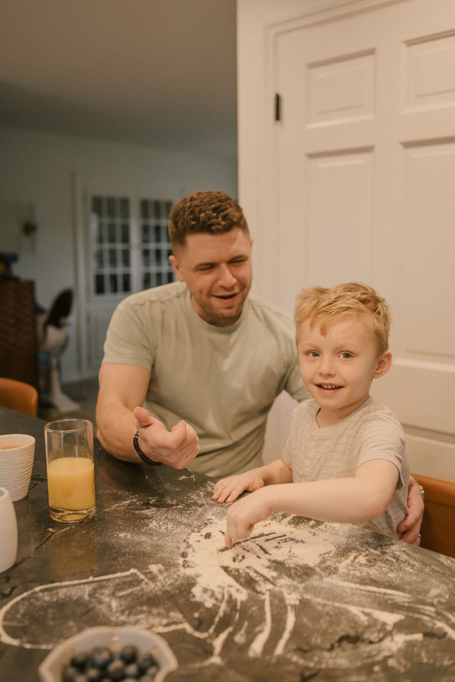 Father and young red-haired son smile together at a flour-dusted kitchen counter with orange juice.