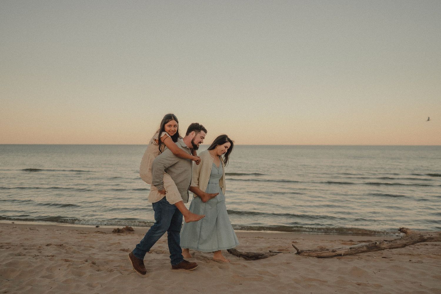Family of three walks along sandy beach at sunset with calm ocean in background.
