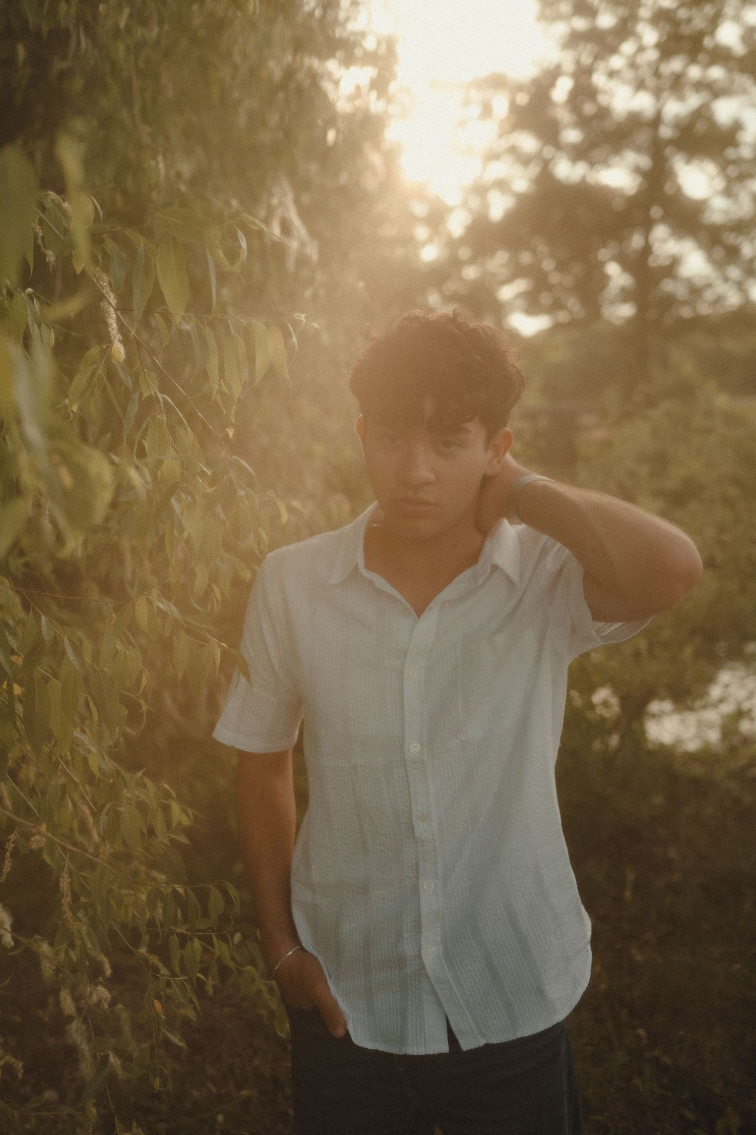 Young man in white linen shirt poses outdoors during golden hour sunset surrounded by lush green foliage.