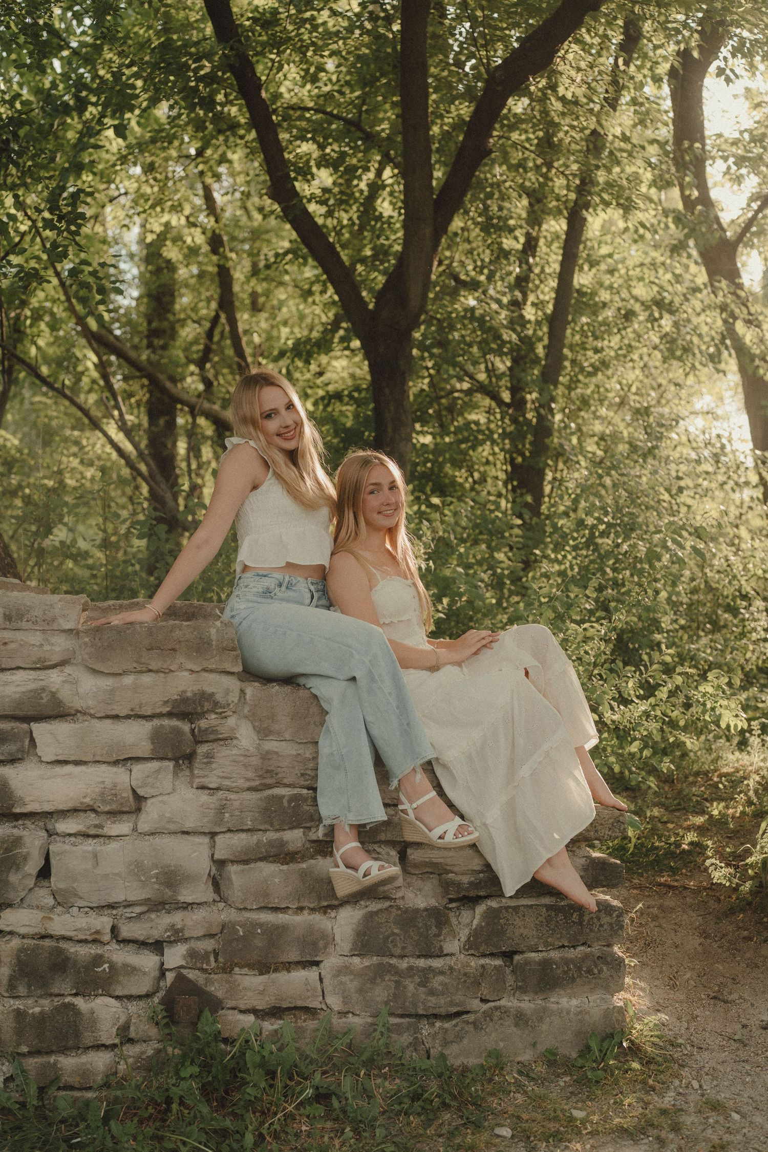 Two young women sitting on a rustic stone wall surrounded by lush green trees in a sunlit outdoor setting.