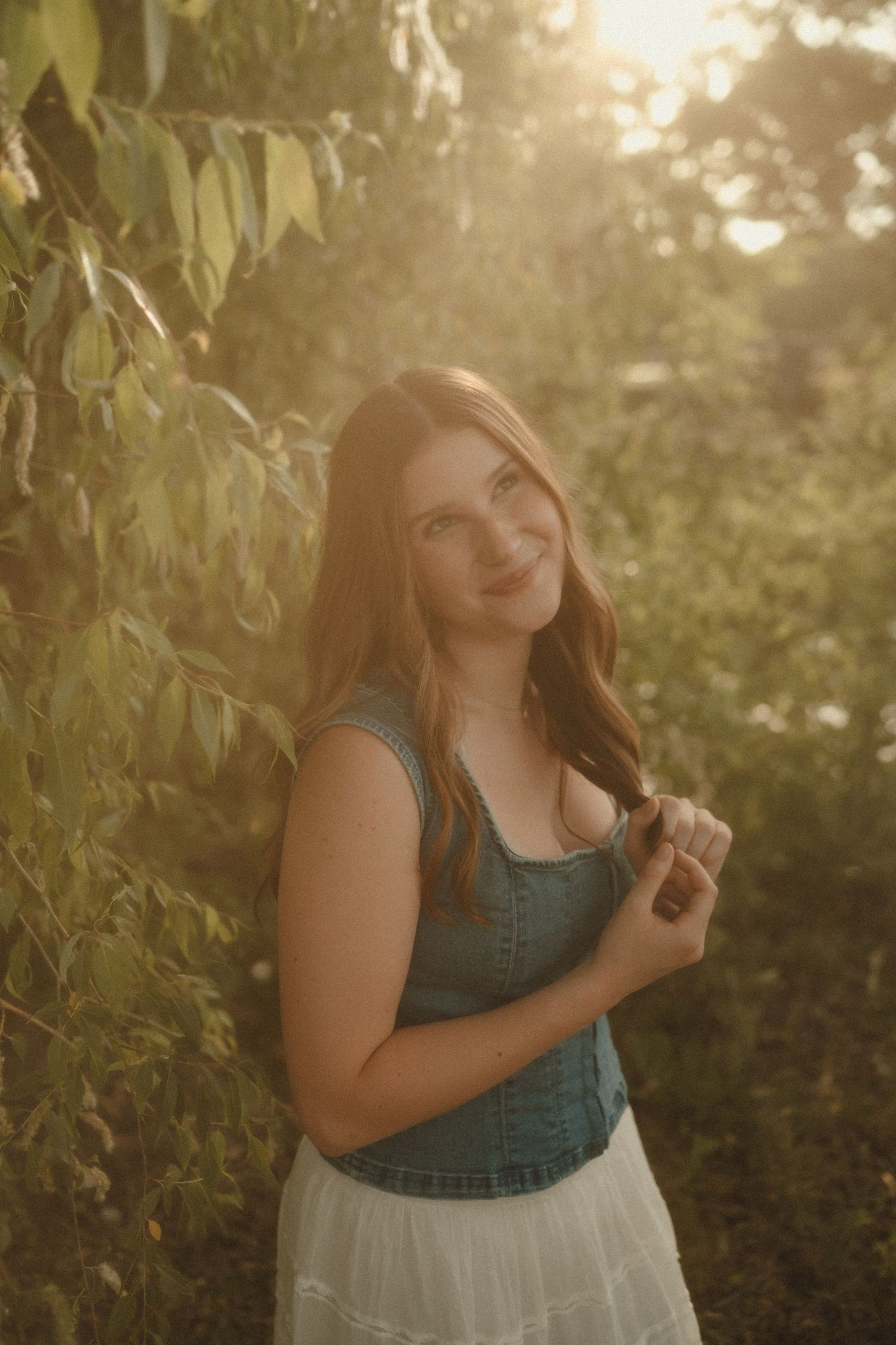 Young woman in denim top and white skirt smiling outdoors in golden sunlight among lush green trees.