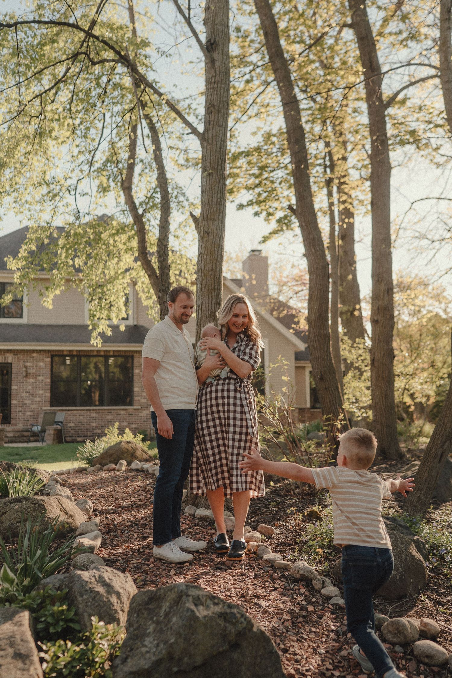 Family of four enjoying a sunny day outdoors near a stone garden with tall trees and a house in the background.