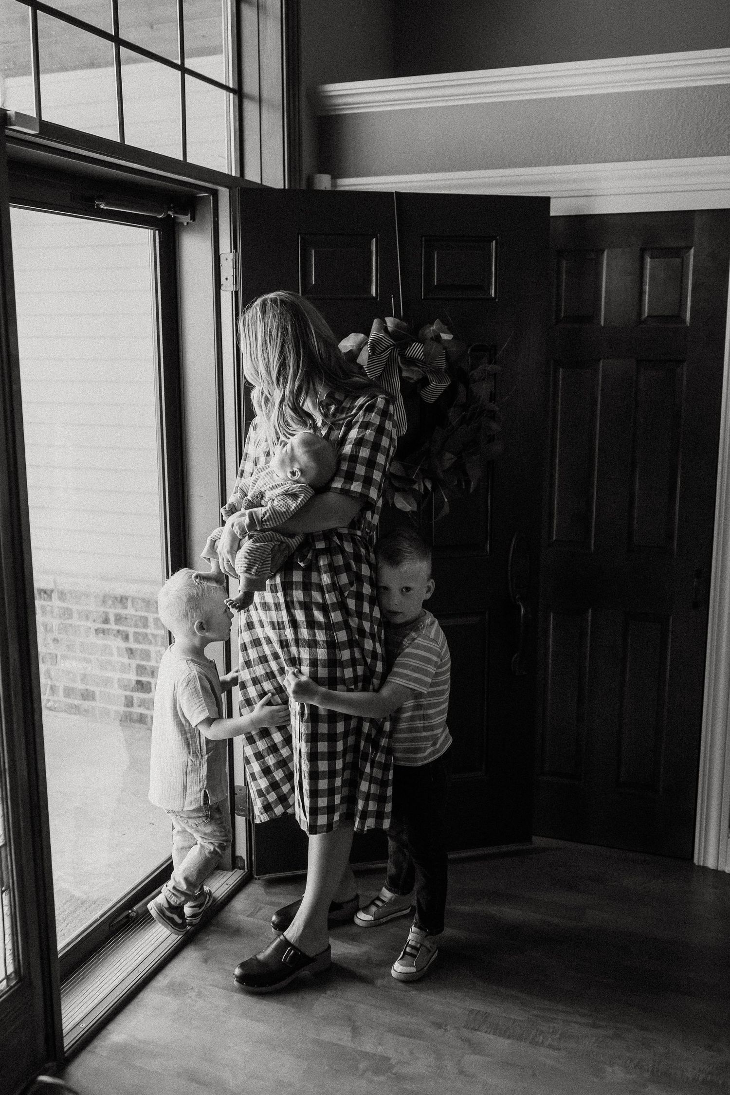 Black and white photo of a mother holding her baby near a front door while two young children hug her.