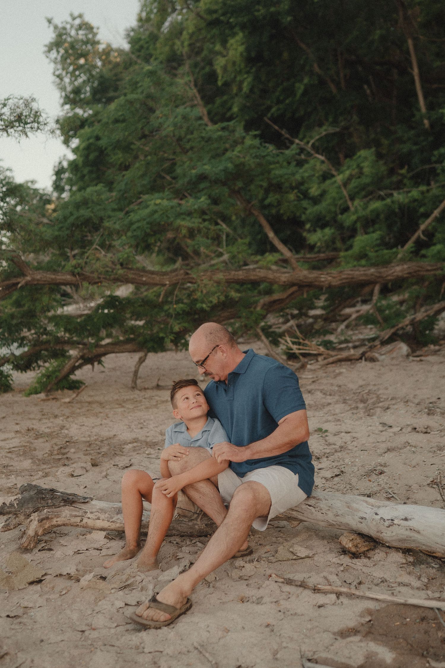 Father and son sharing a quiet moment sitting together on a sandy beach near trees.