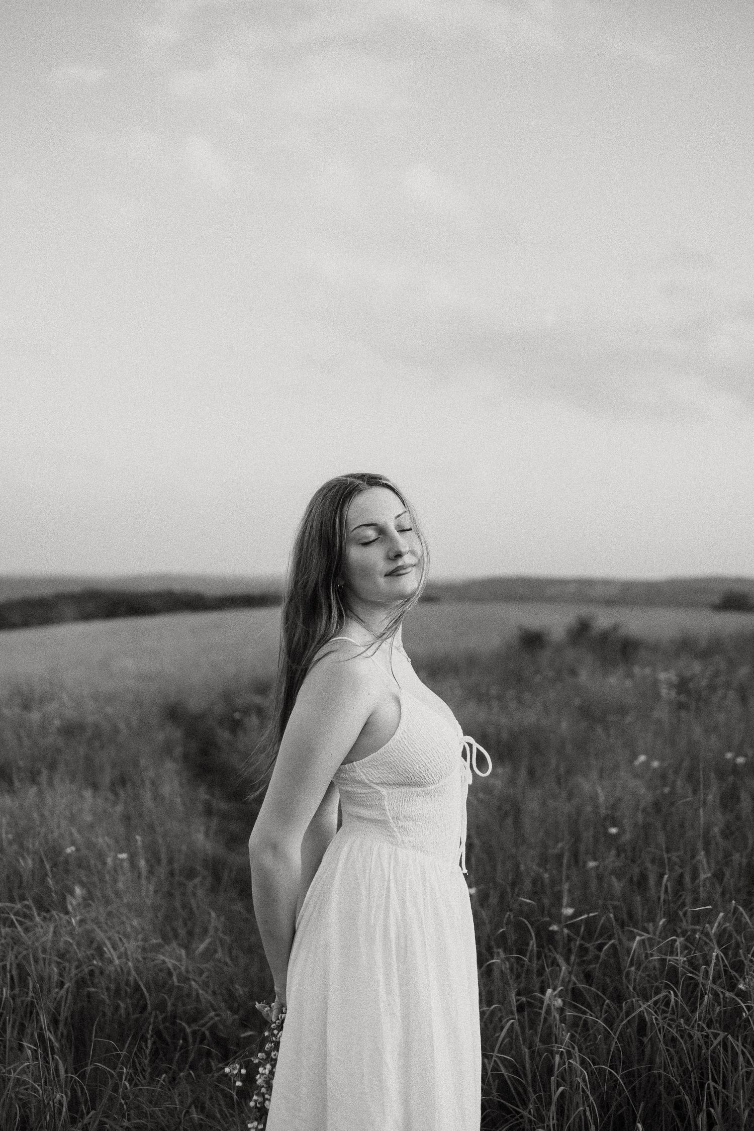 Black and white portrait of young woman in white dress standing in field with eyes closed, serene expression.