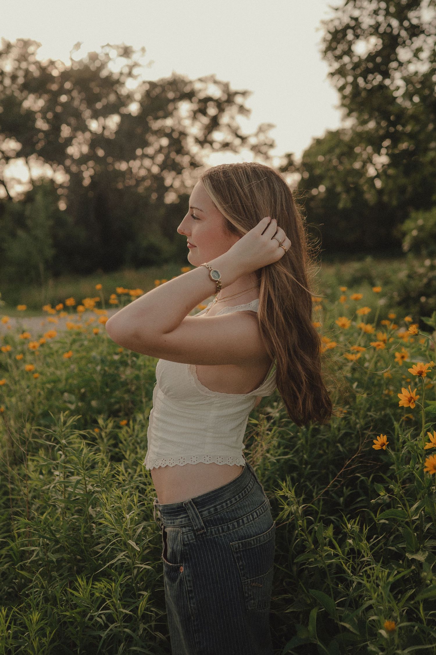 Young woman in white crop top and jeans poses among yellow wildflowers at golden hour sunset.