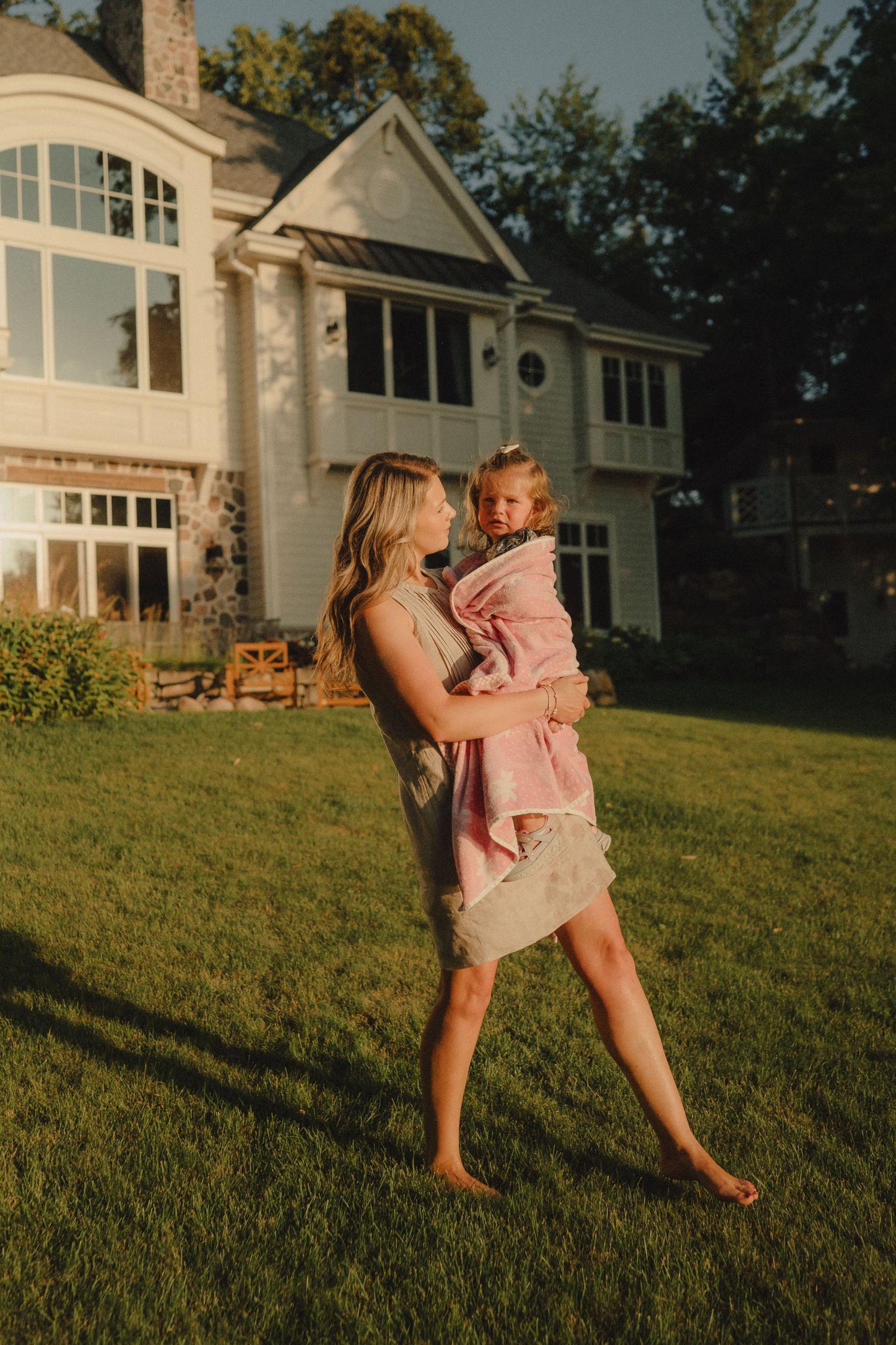 A barefoot mother carries her toddler wrapped in a pink towel outside a large lakeside home.