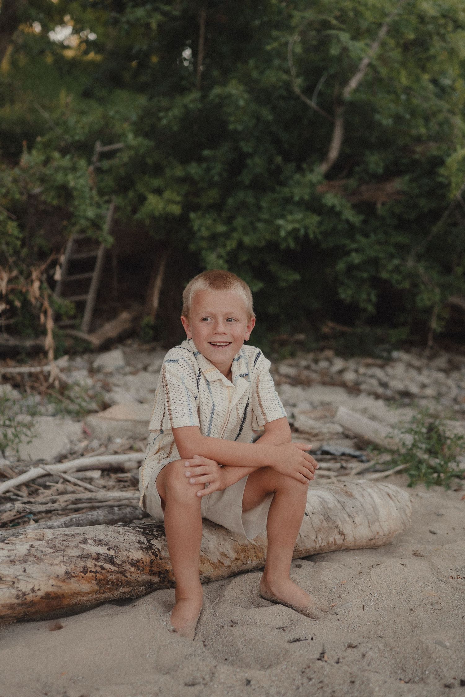 Happy young boy sits cross-legged on driftwood on a sandy beach with lush trees.