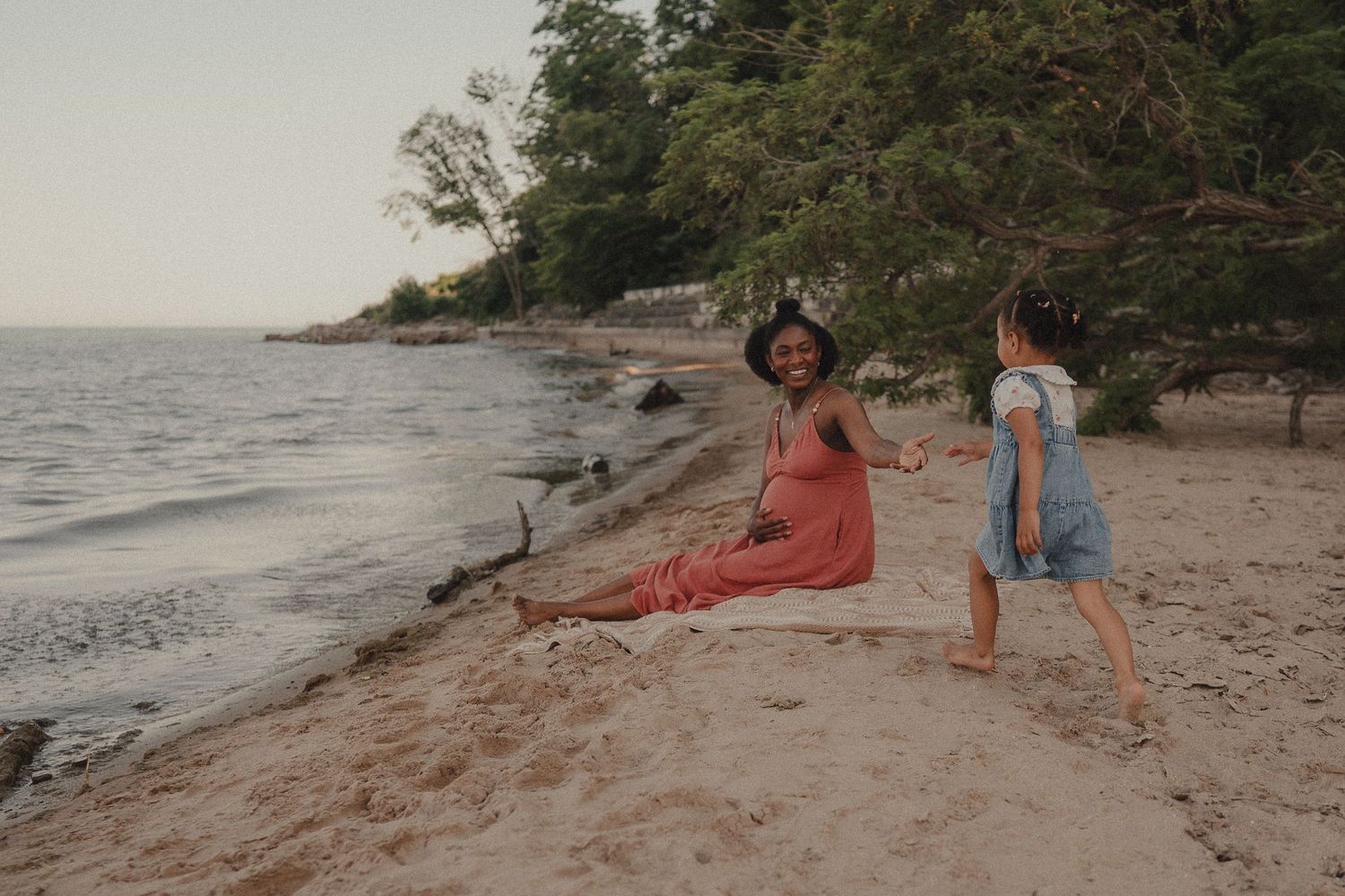 Woman in red dress sits on sandy beach while child in blue dress stands nearby, with trees and calm water in background.