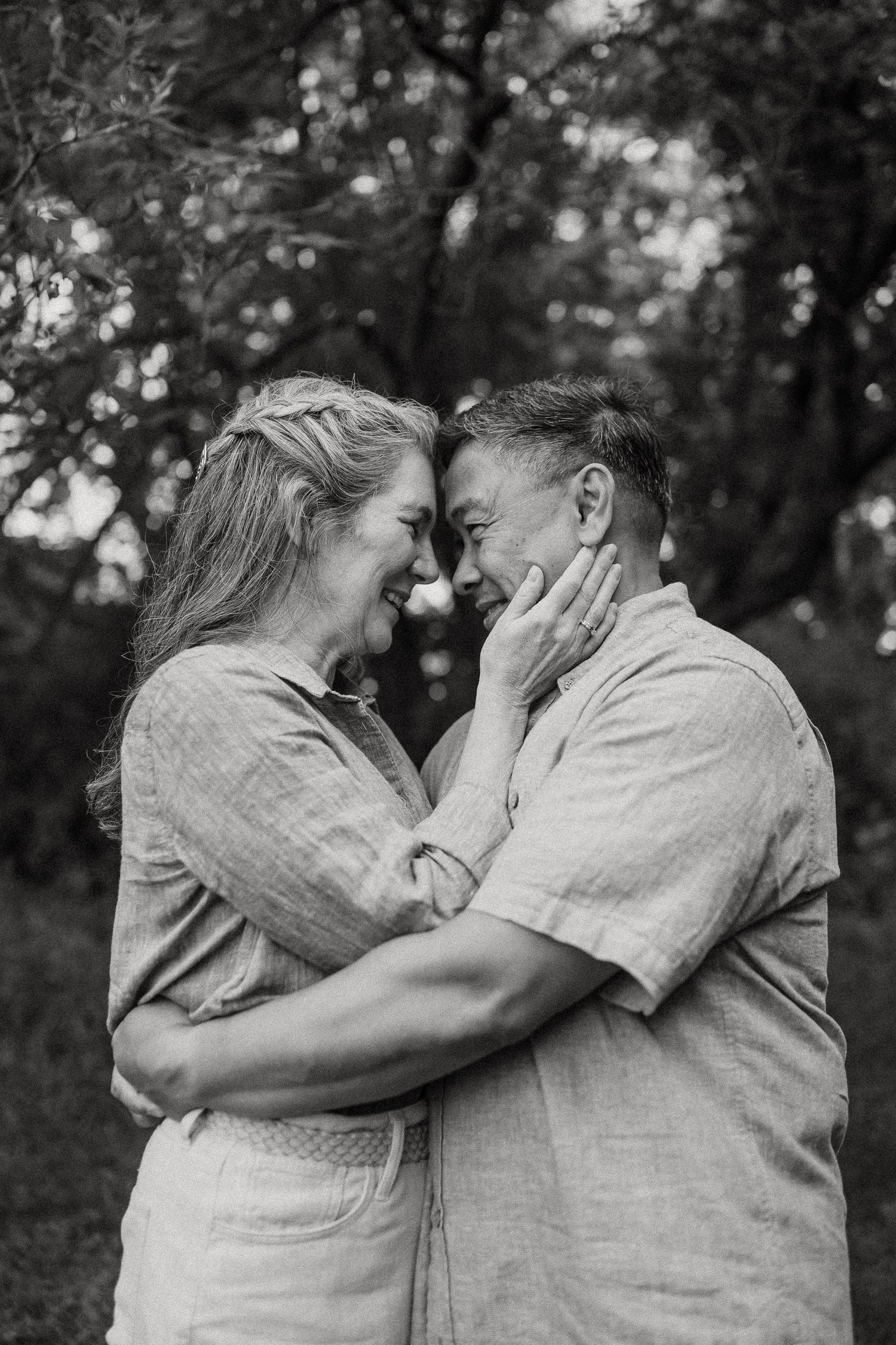 Black and white photo of a loving couple touching foreheads outdoors, woman gently cupping mans face among trees.
