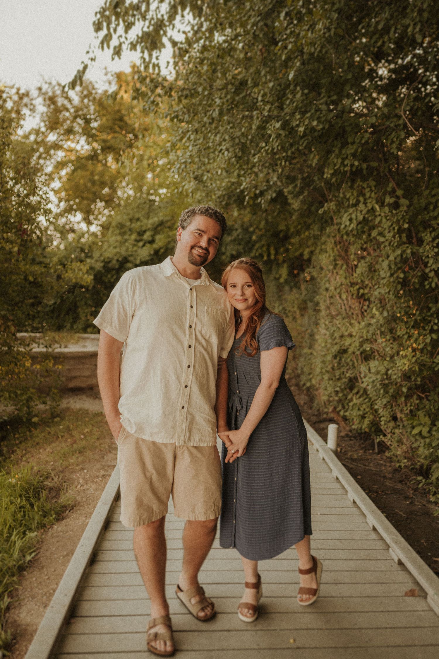 Couple poses together on a wooden boardwalk path surrounded by green foliage in warm sunlight.