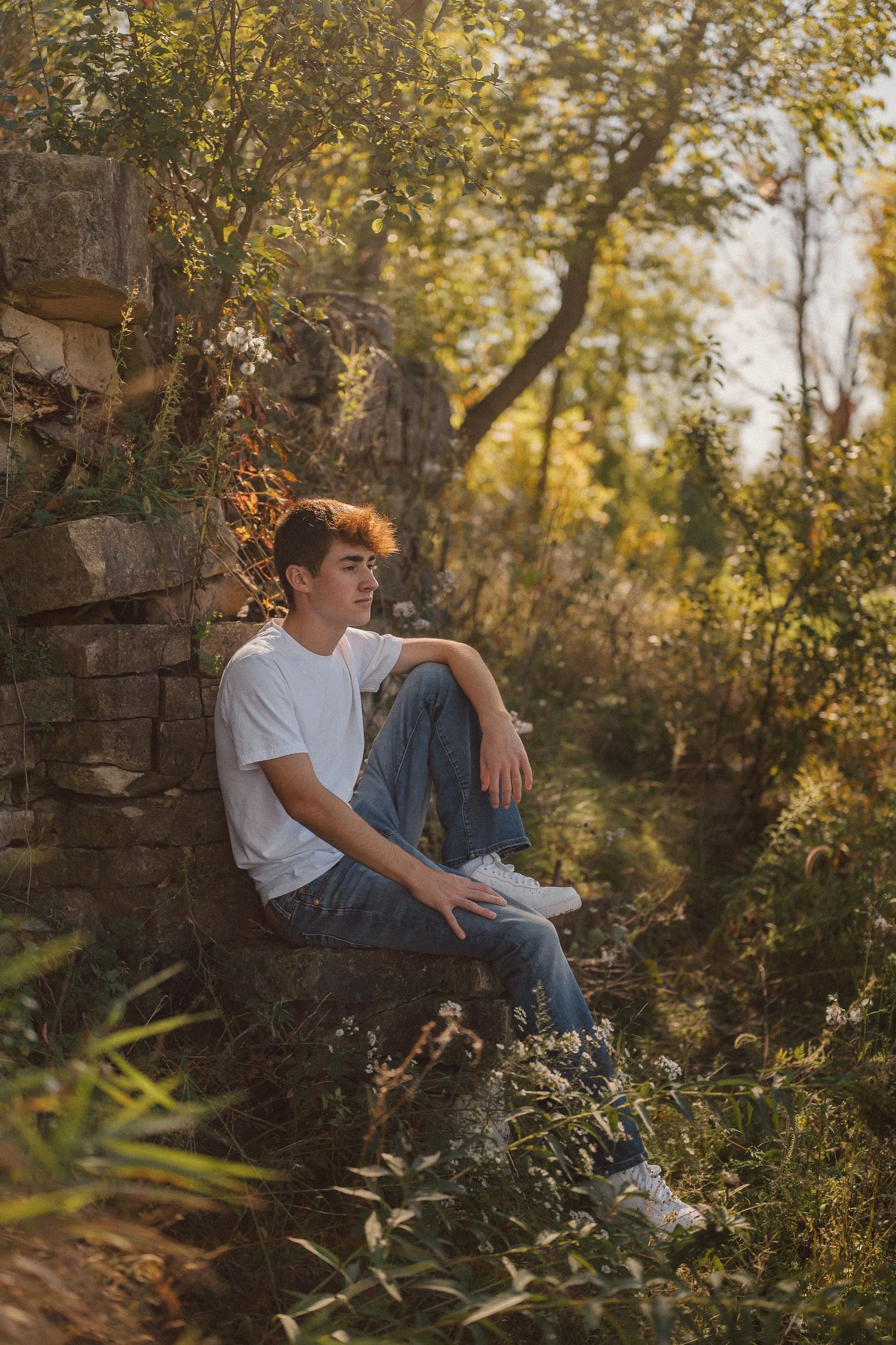 Person in white t-shirt and jeans sits casually against stone wall surrounded by autumn foliage in natural setting.
