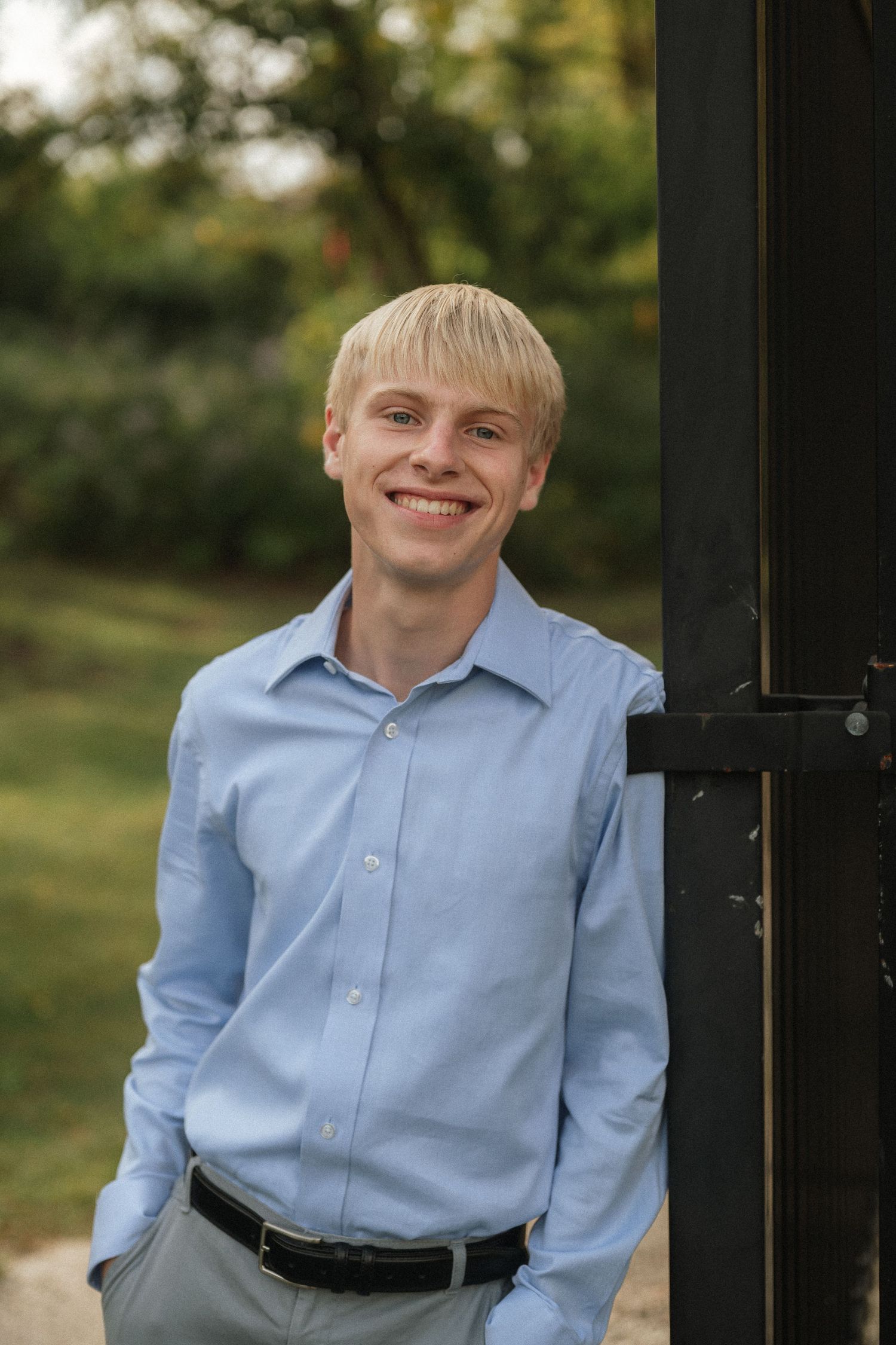 A person in a light blue button-down shirt smiles while standing against a dark post outdoors.