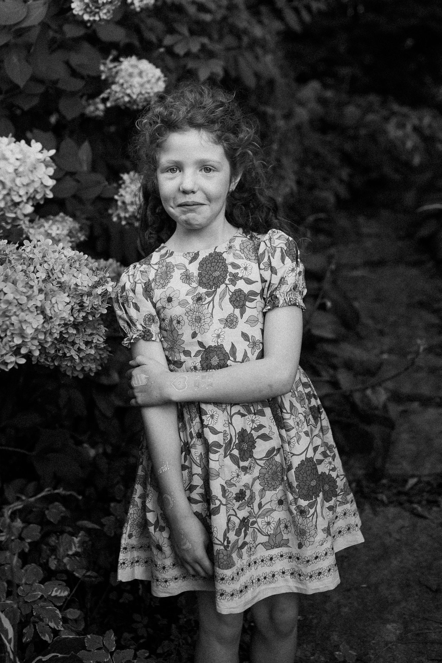 A child in a floral dress stands against a dark background in a black and white photograph.