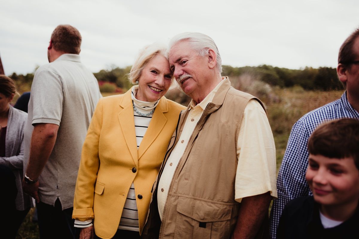 An affectionate elderly couple embracing outdoors, with the woman wearing a yellow blazer and the man in a tan vest.