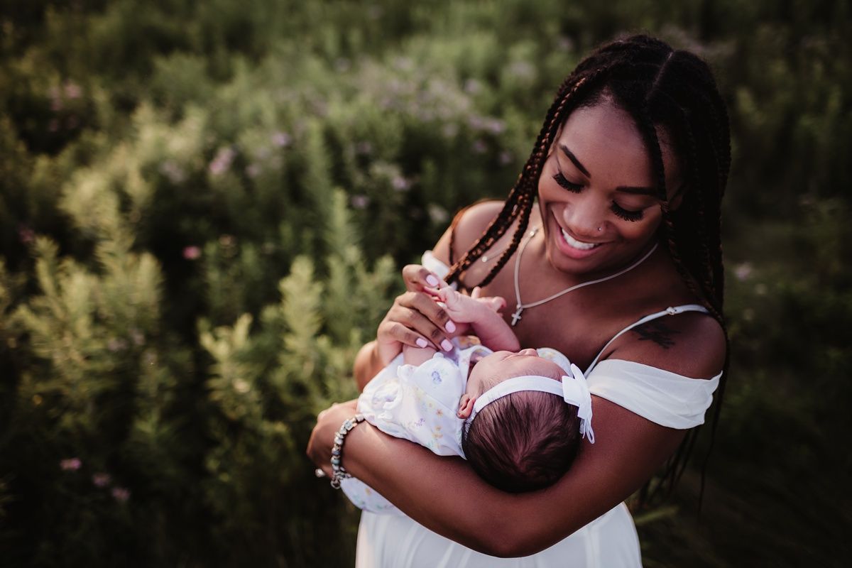 A woman in an off-shoulder white dress smiles while holding a newborn outdoors.