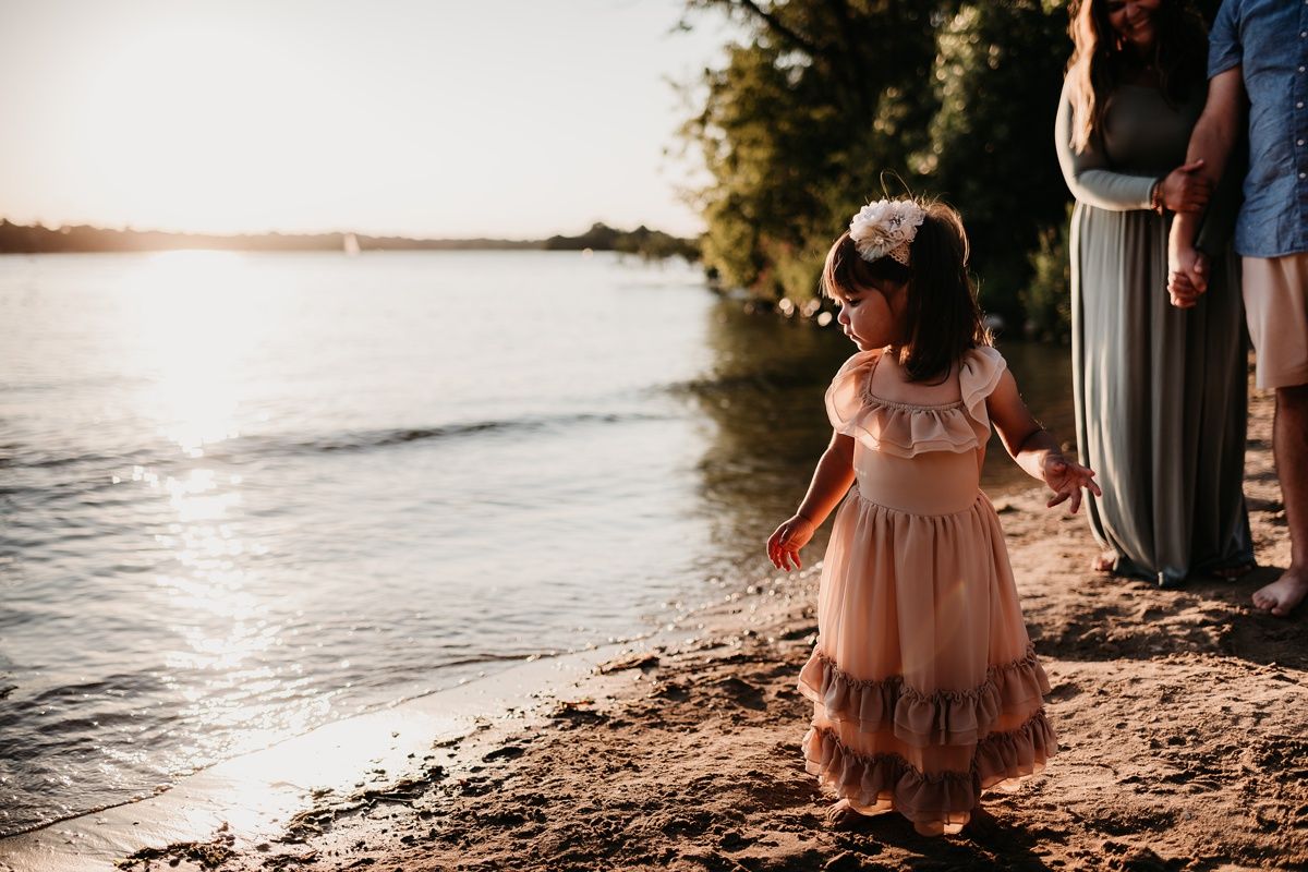 Child in flowing dress stands at water's edge during golden hour.
