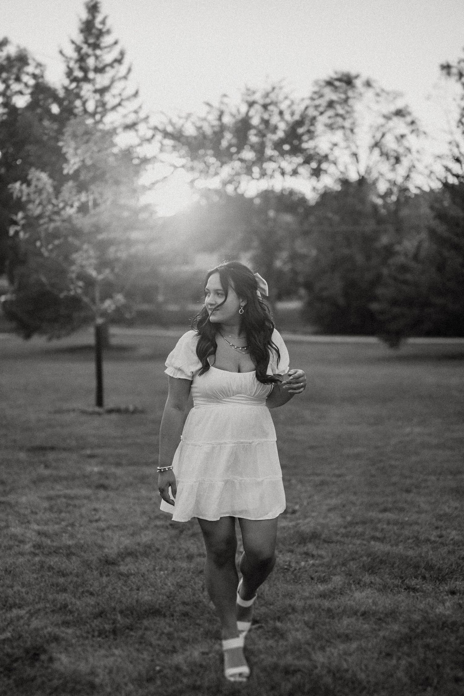 Black and white photo of someone in white dress walking through park at sunset with backlit trees.