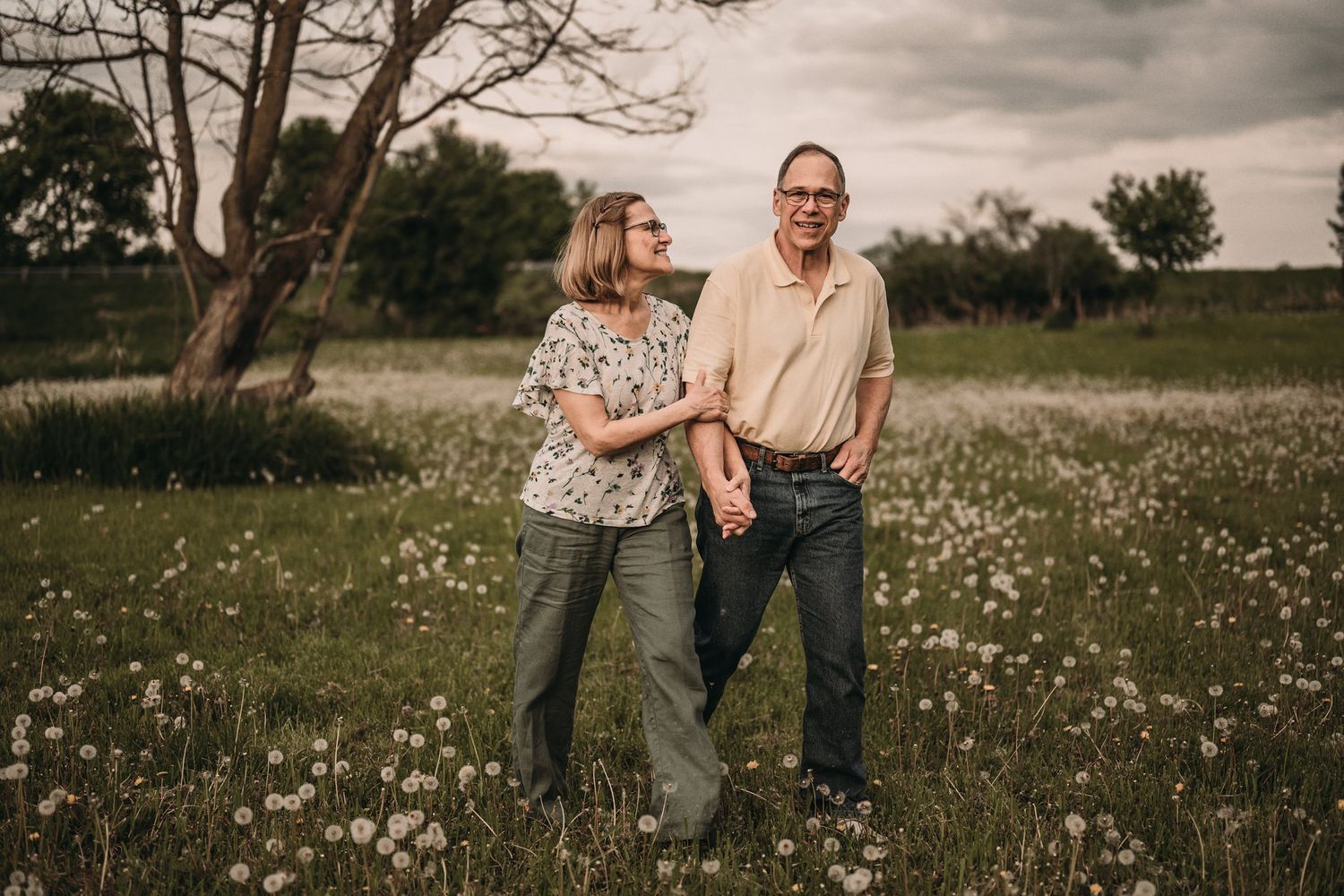 Two people stand together in a flowering meadow at sunset with a tree silhouetted in background.