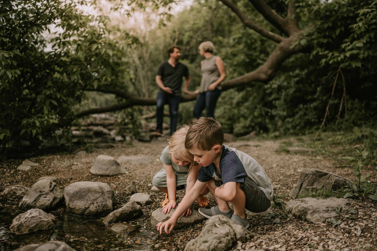 Child exploring rocks and nature while others watch in a wooded outdoor setting.
