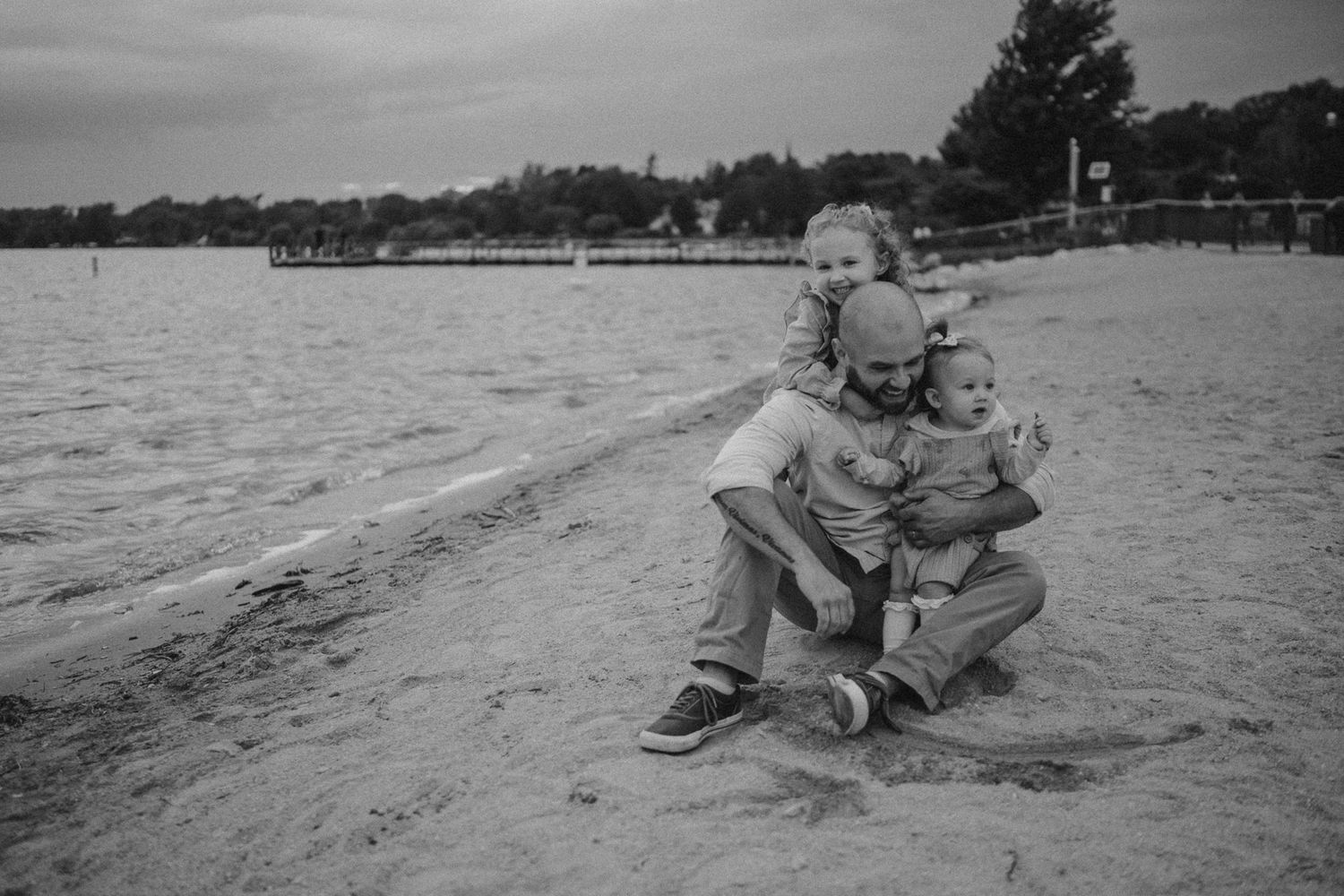 A black and white photo of playful interaction on a lakeside beach with docks in the background.