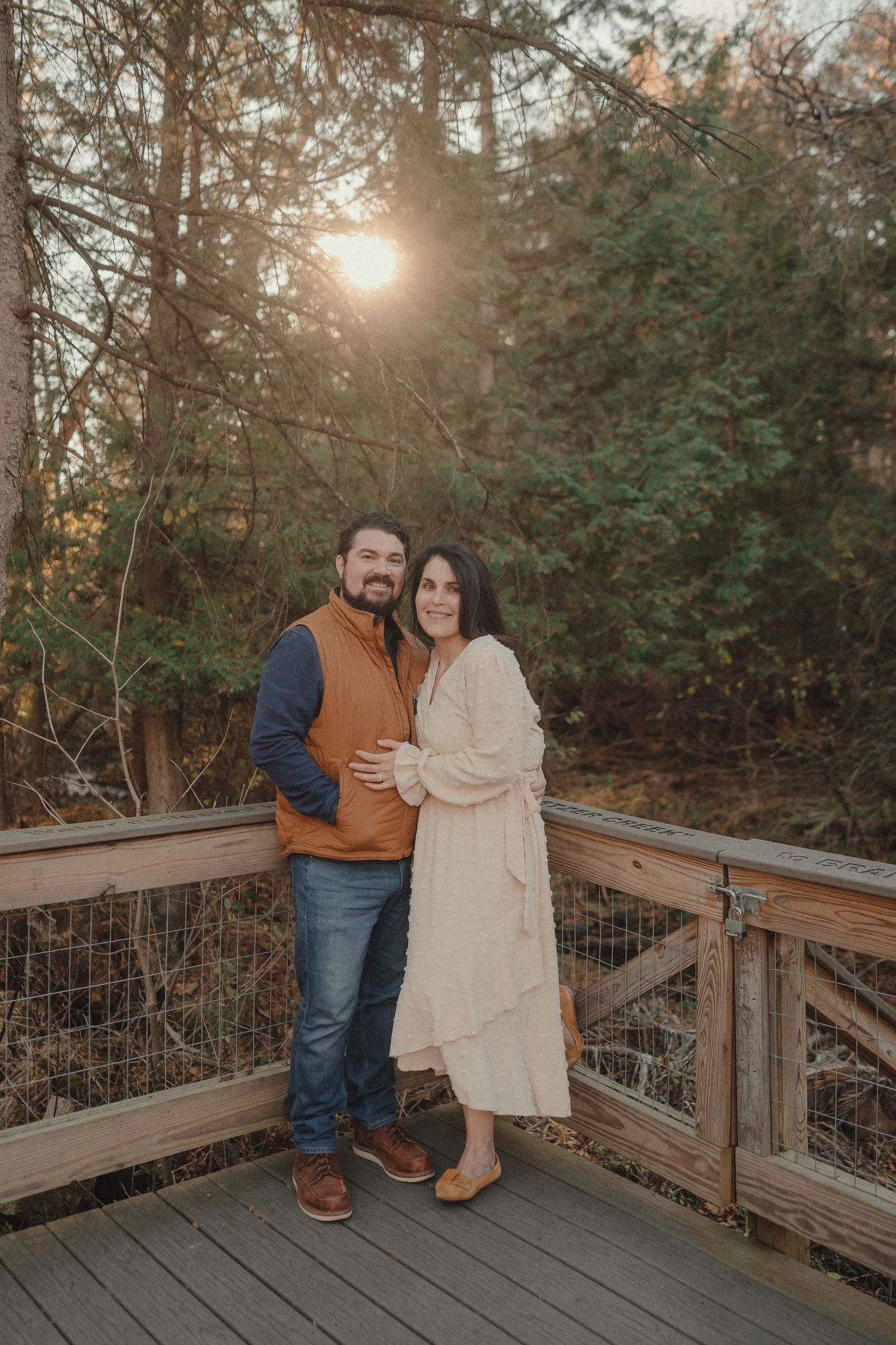 A couple poses together on a rustic wooden deck with forest scenery and sunlight in the background.