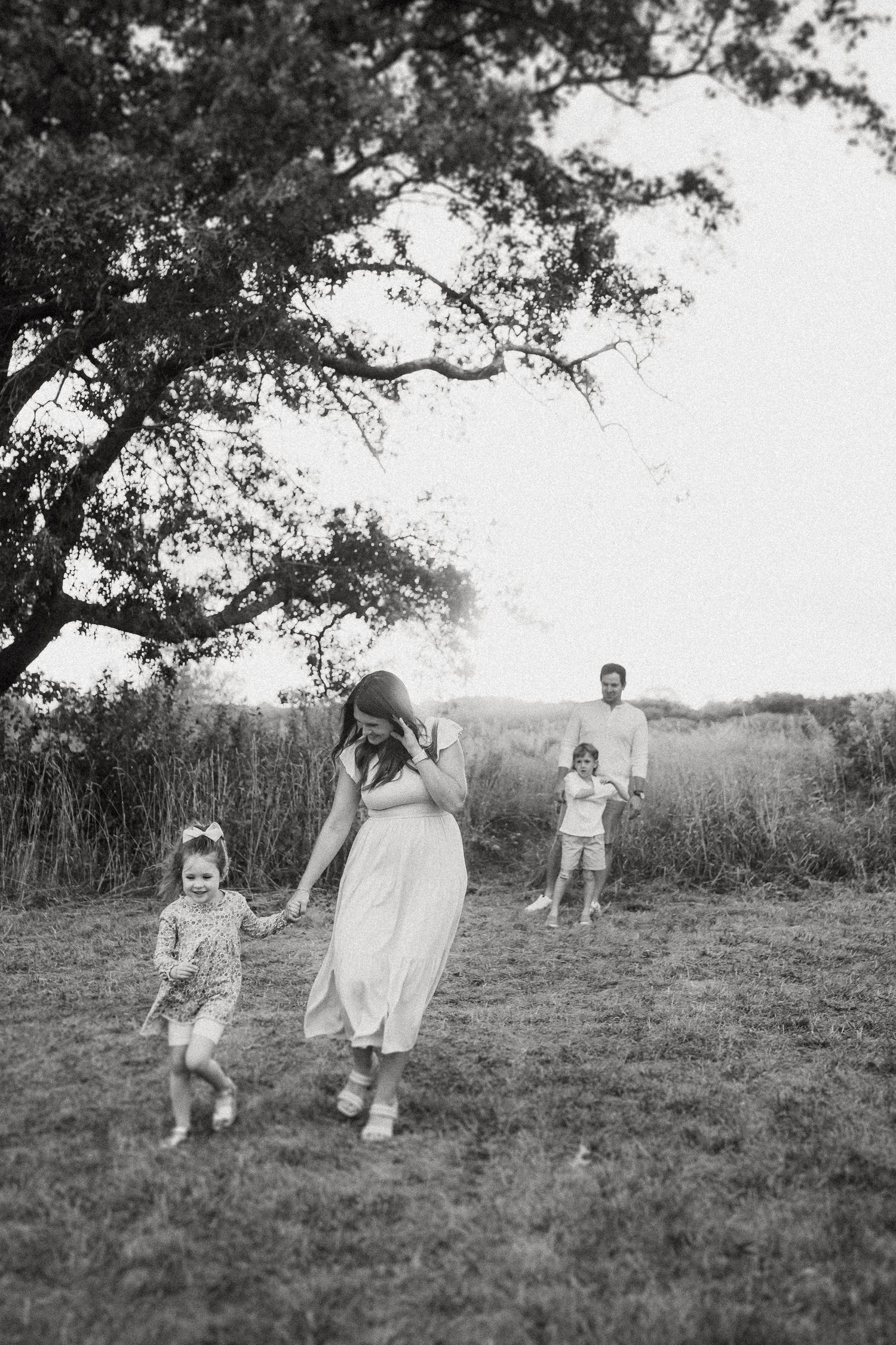 Family walking together in a meadow under large tree branches in black and white.