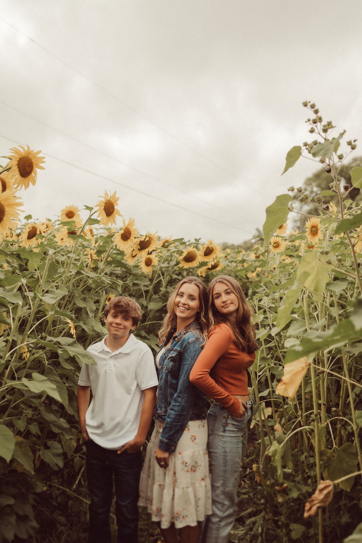 Three people pose together among tall sunflowers on an overcast day.