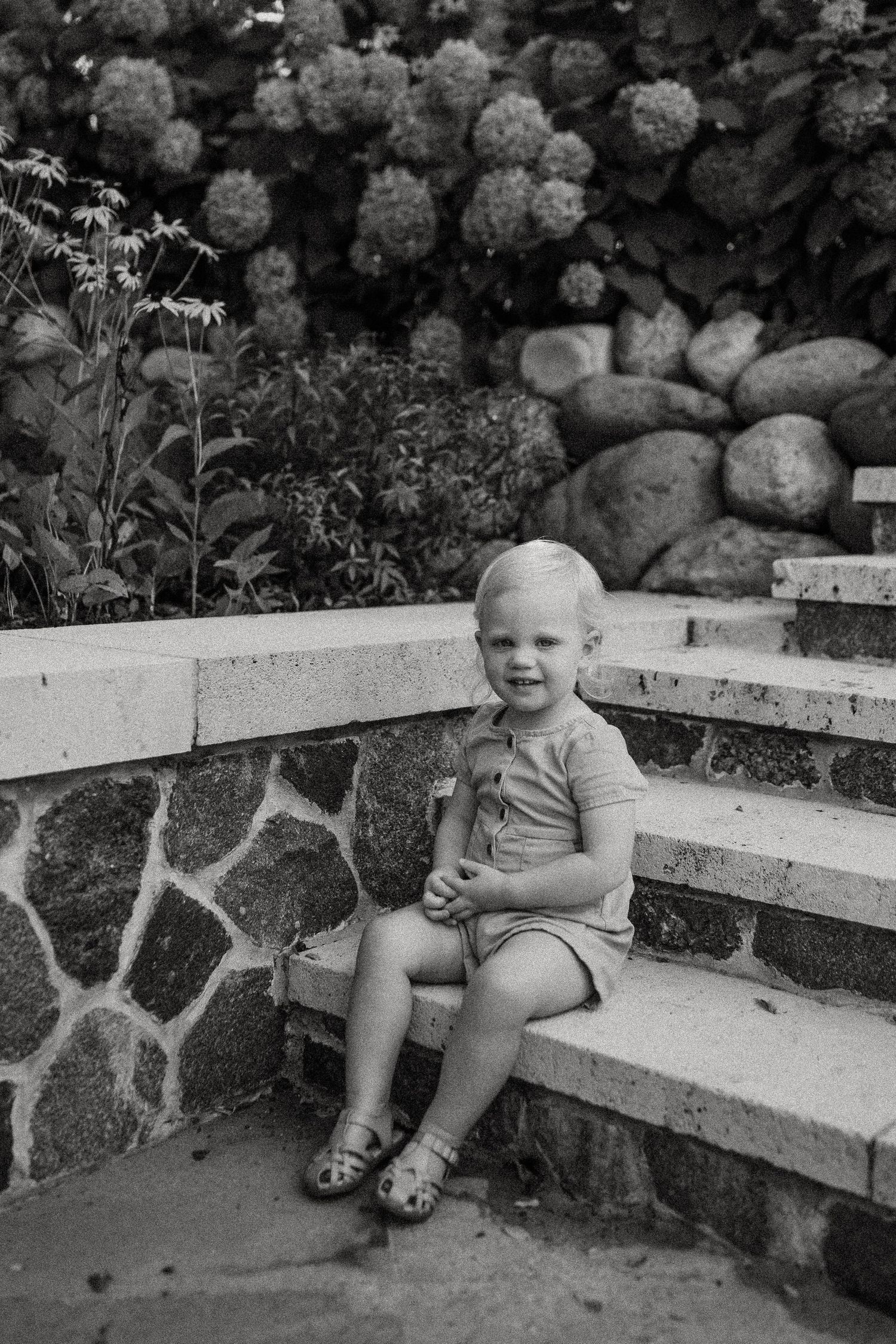 Black and white portrait on garden steps with stone retaining wall and flowering bushes.