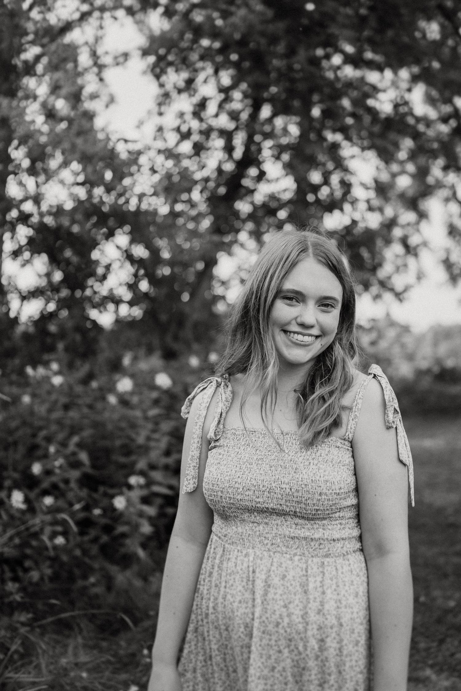 Black and white portrait featuring a smiling figure in a patterned summer dress outdoors.