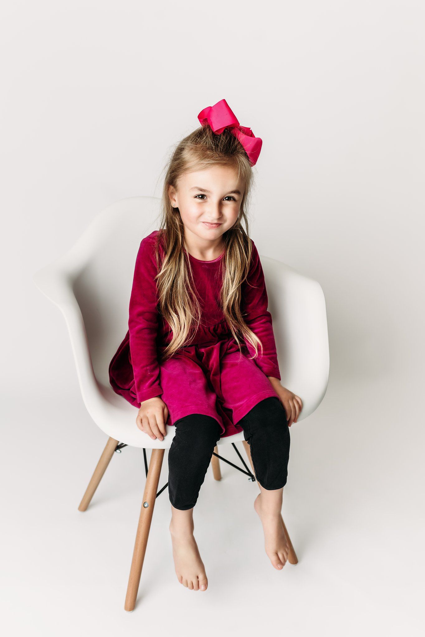 Child in burgundy dress sitting casually on modern white chair.