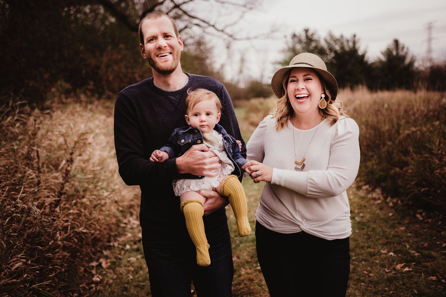Joyful outdoor family portrait with baby in yellow pants against autumn landscape.