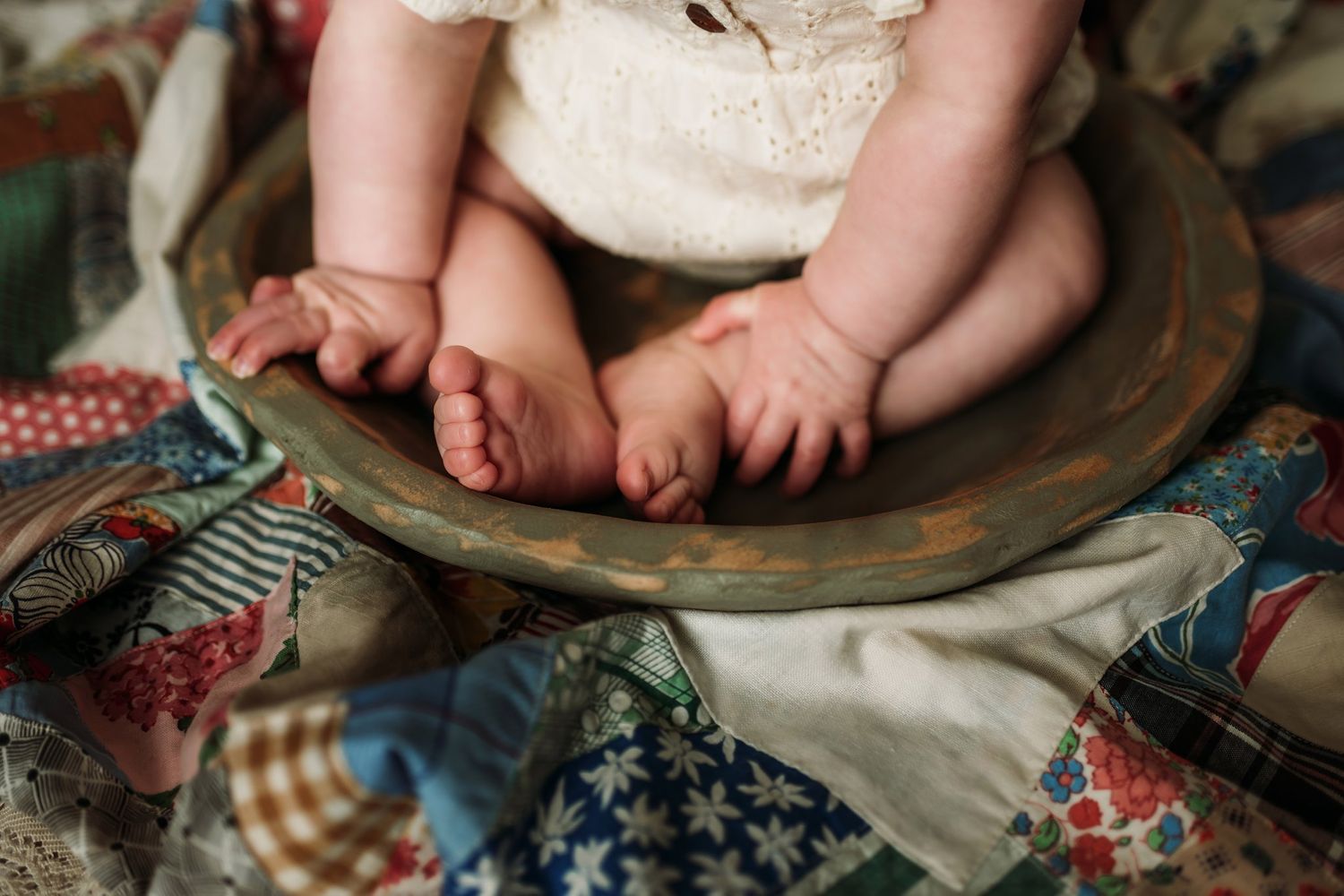 Close up of baby feet and hands resting on wooden bowl with vintage quilt pieces underneath.
