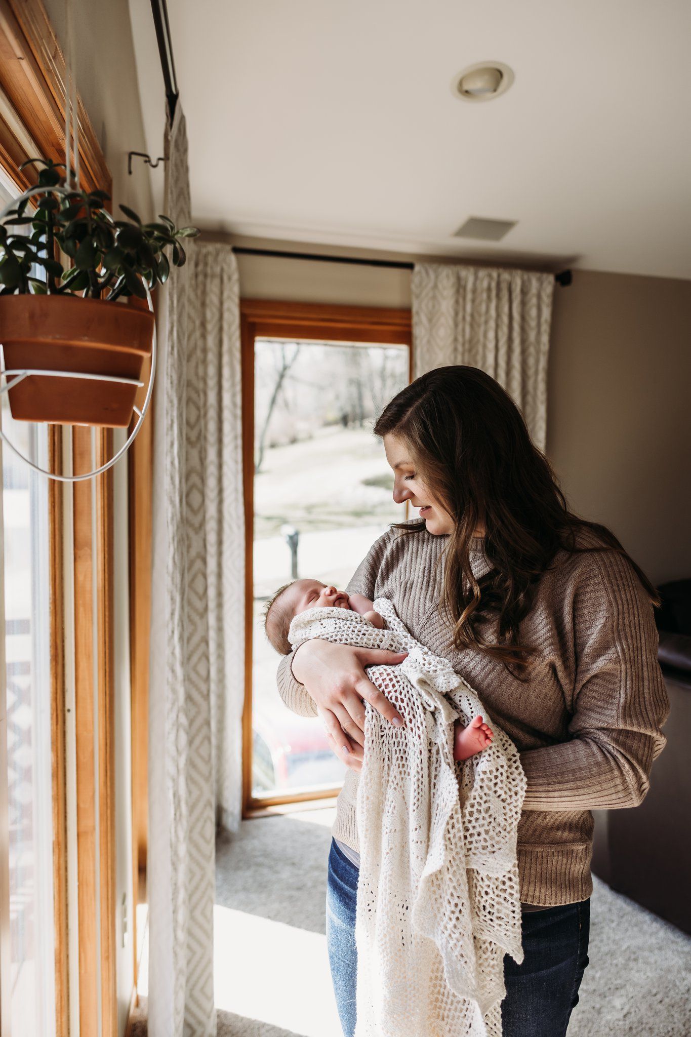 Sunlit window scene with baby wrapped in knit blanket beside hanging plant.