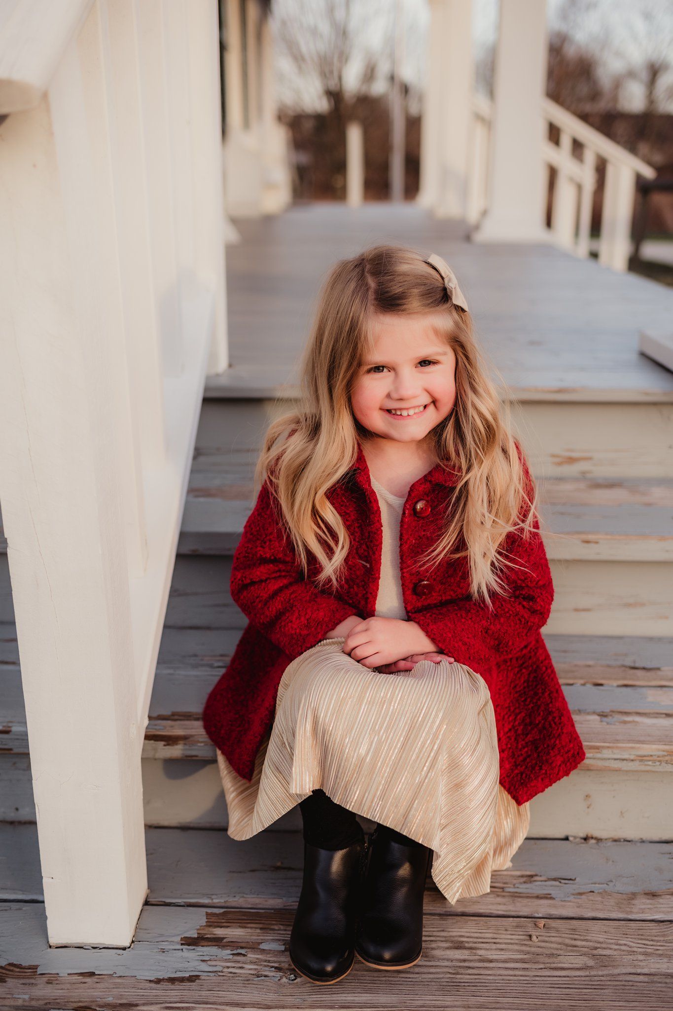 A child in a bright red coat and cream dress sits on wooden porch stairs during a photo session.