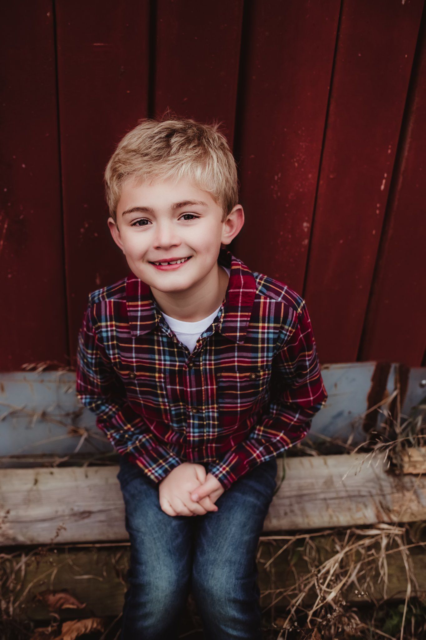 A young boy in a plaid shirt smiles brightly while sitting against a red barn wall.