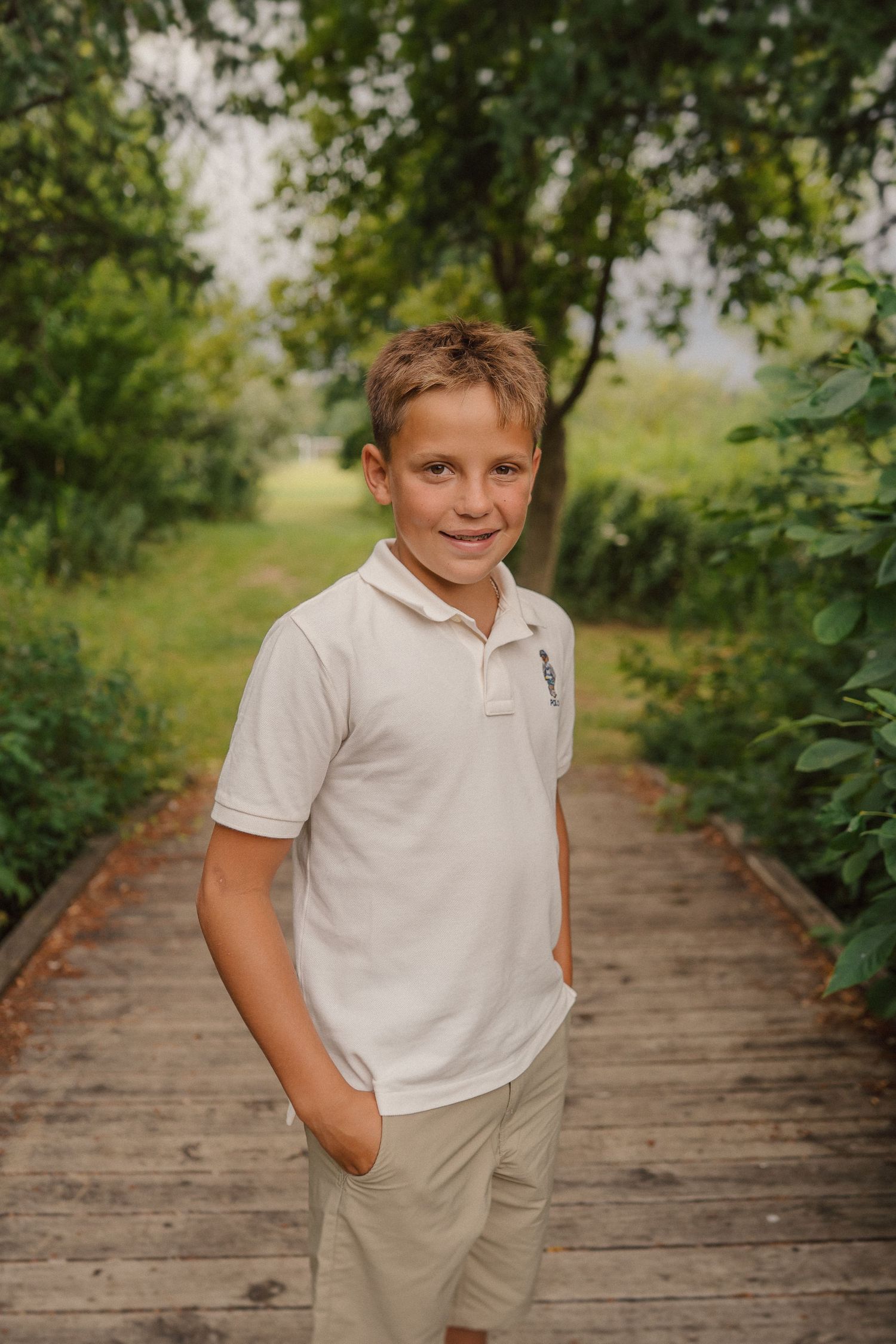 Young boy in light colored polo and khaki shorts standing casually on wooden boardwalk.