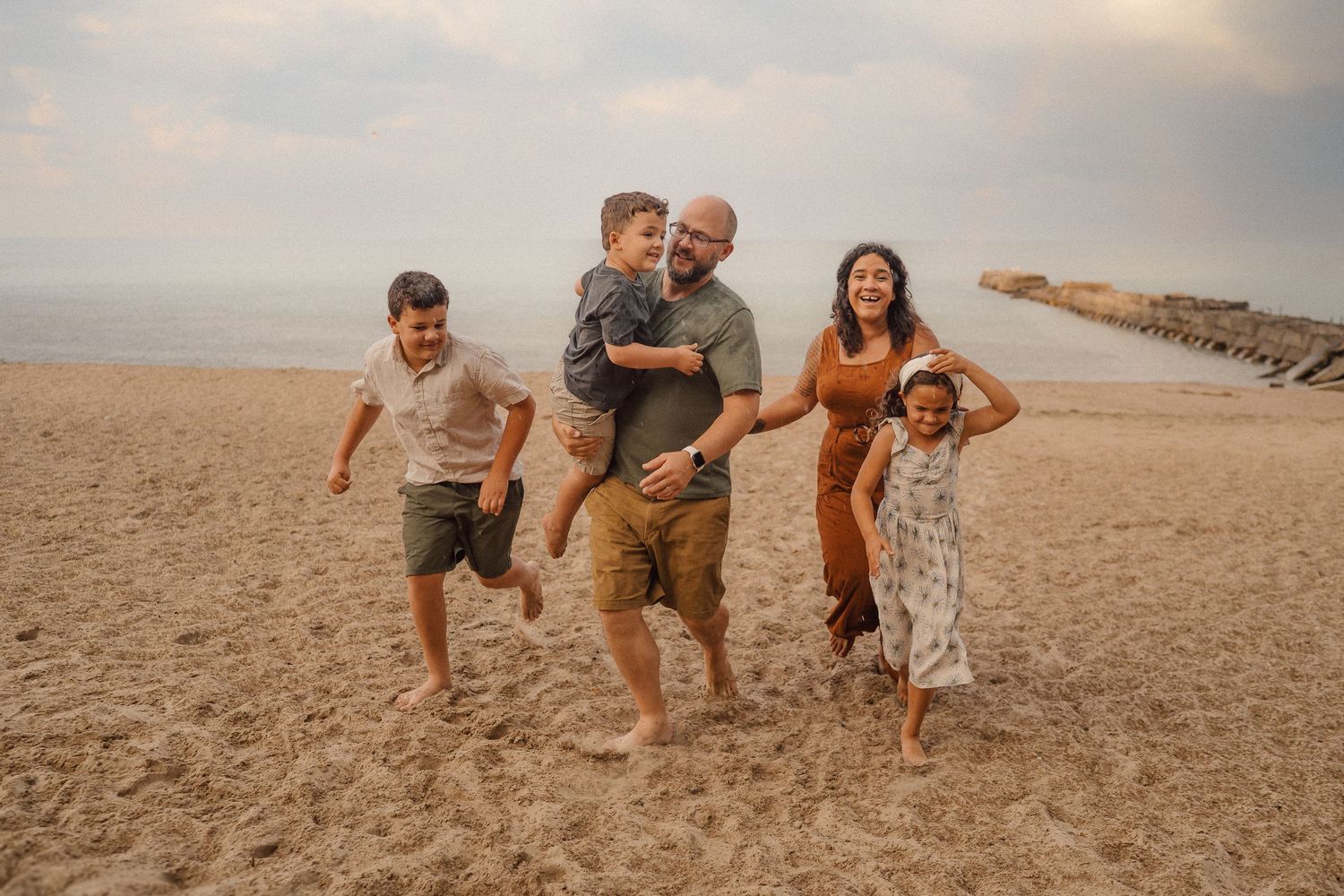 A playful family runs and jumps together on a sandy beach during golden hour with cloudy skies.