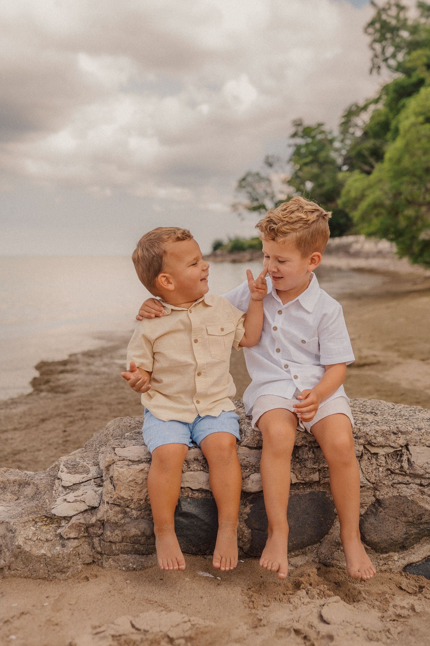 Two children in casual summer outfits sit together and laugh on rocky beach during cloudy day.