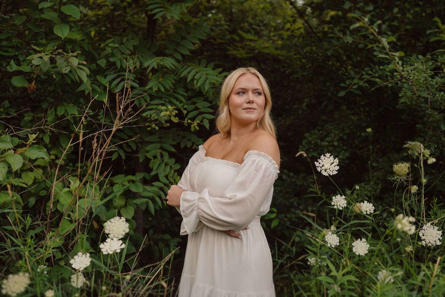 A person in a white flowy wedding dress poses in front of lush green foliage and white flowers in a garden setting.