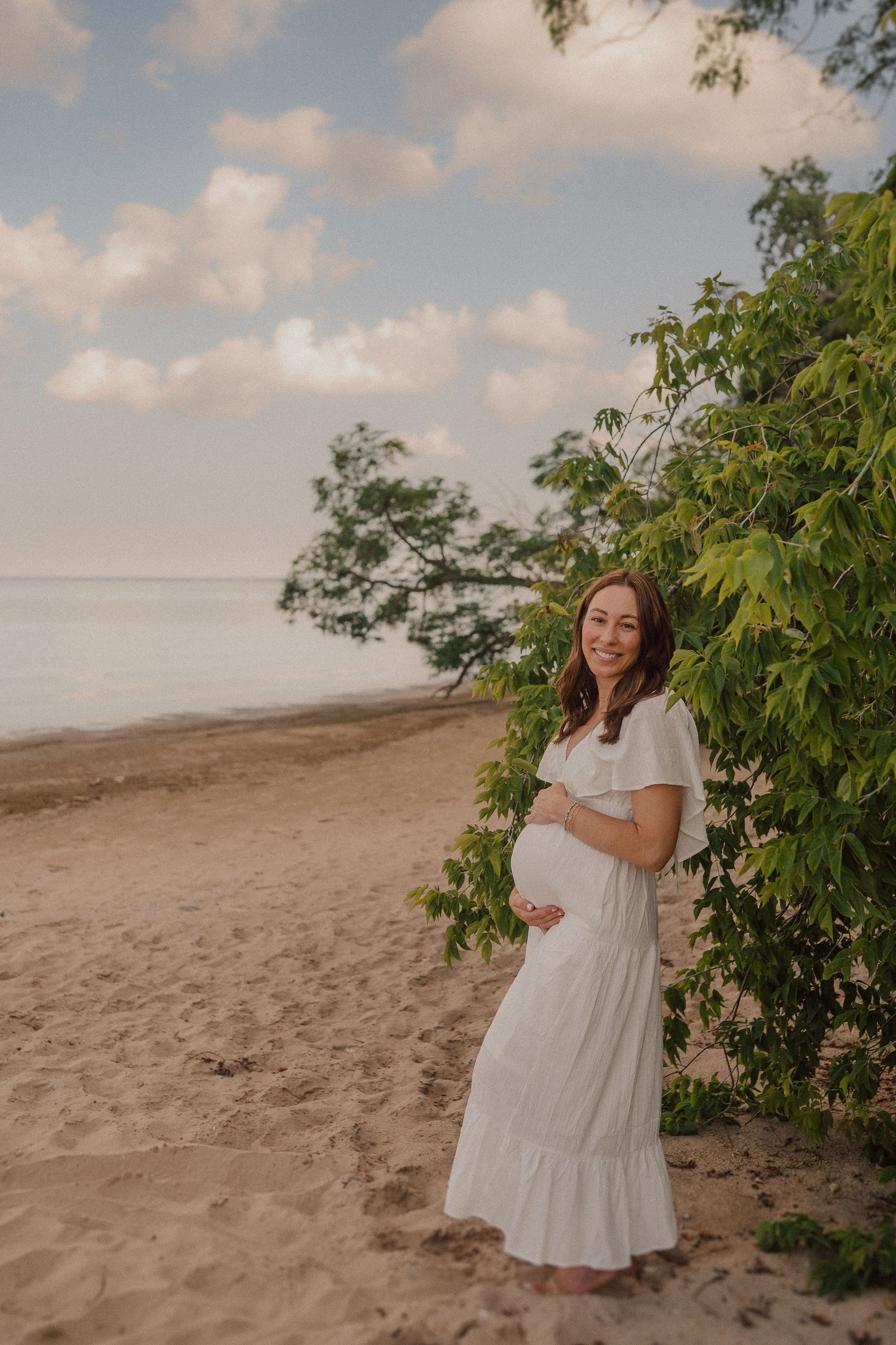 Woman in white dress smiling during maternity photoshoot on sandy beach with lush foliage backdrop.