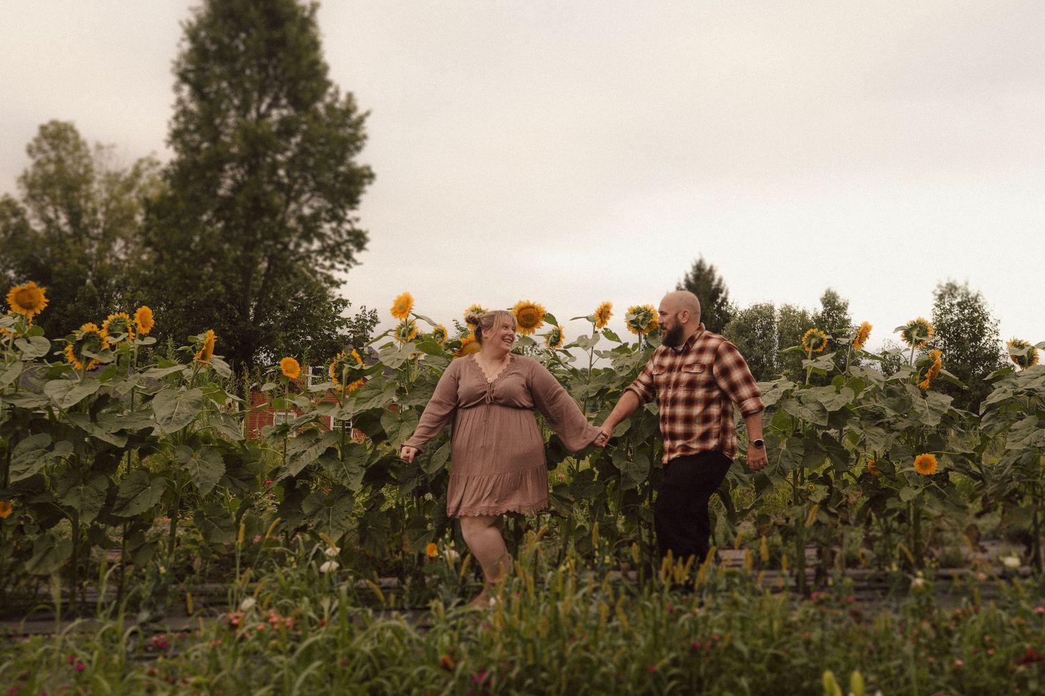 A loving couple holds hands while walking through a lush garden of wildflowers and tall plants.